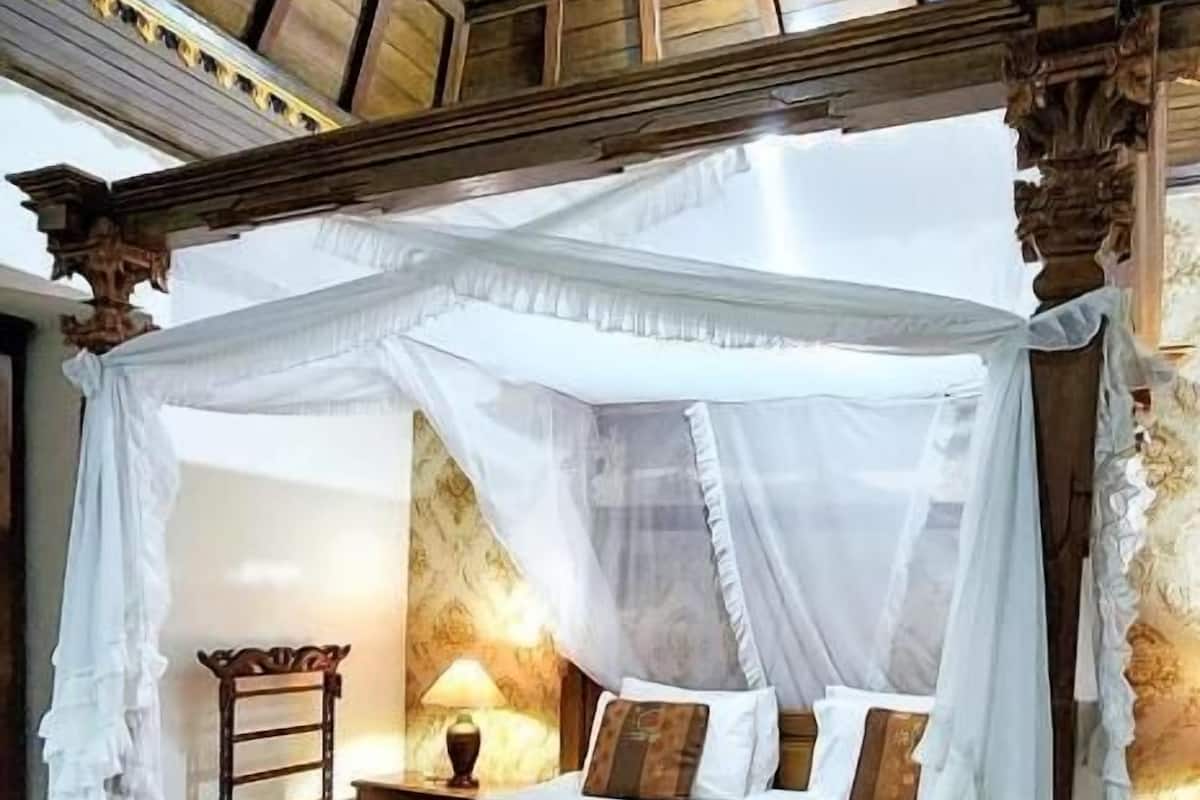 A four-poster canopy bed with a red runner, a nightstand with a lamp, and a view of the wooden ceiling structure.