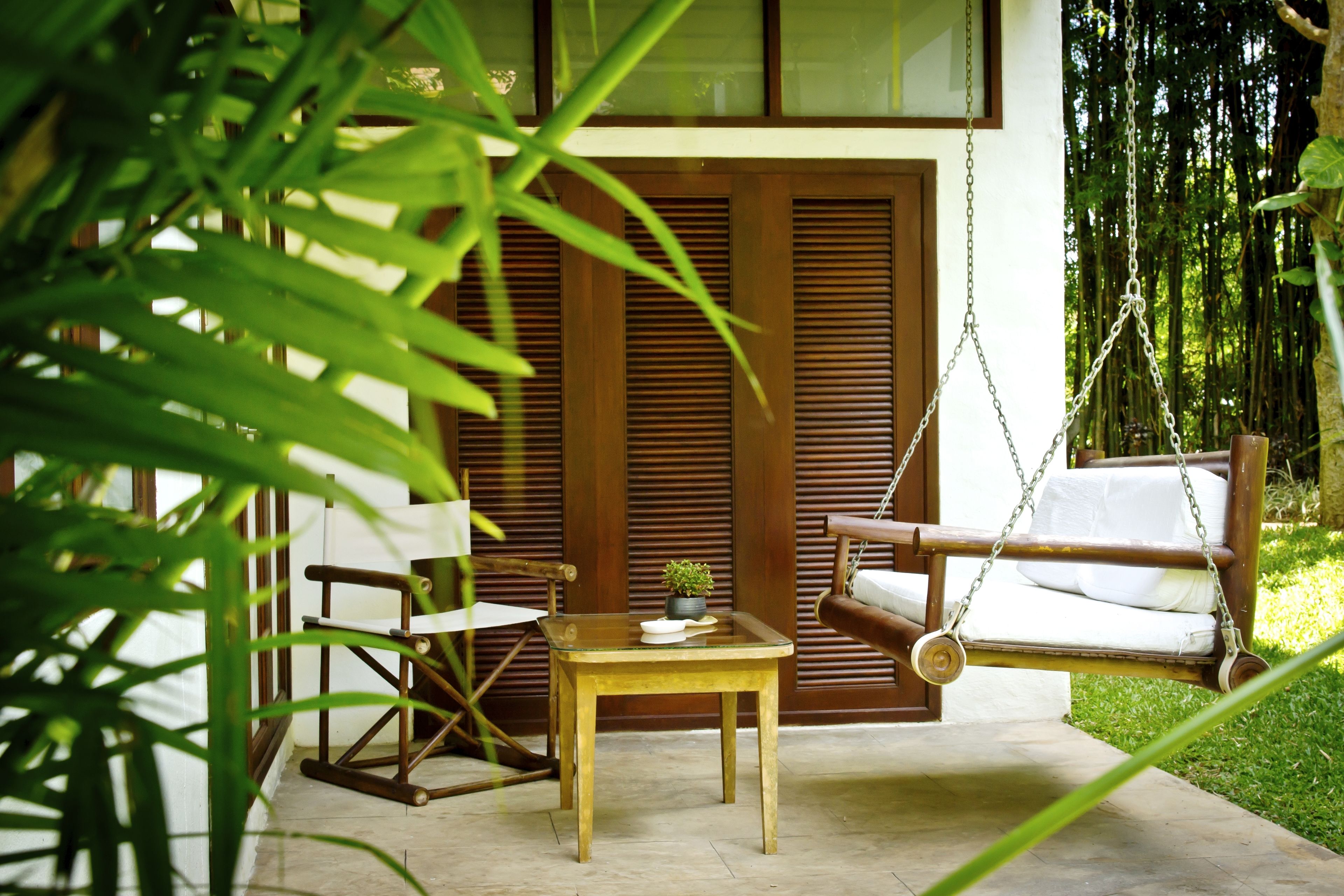 A wooden swing chair, a small table with a plant, and a wooden chair in a garden area with a bamboo backdrop.