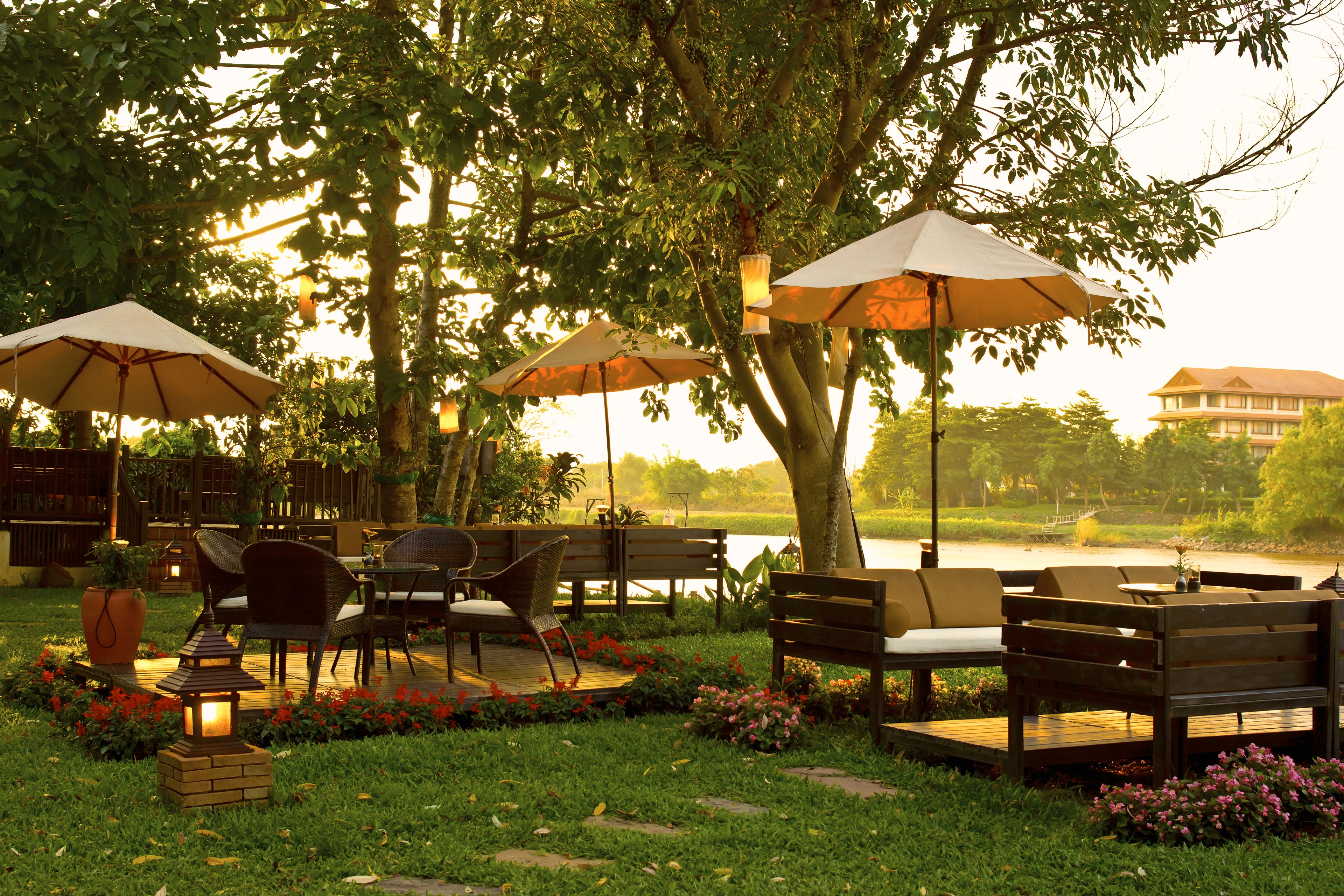An outdoor seating area with tables and chairs under umbrellas, surrounded by trees and a body of water.