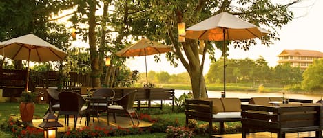 An outdoor seating area with tables and chairs under umbrellas, surrounded by trees and a body of water.