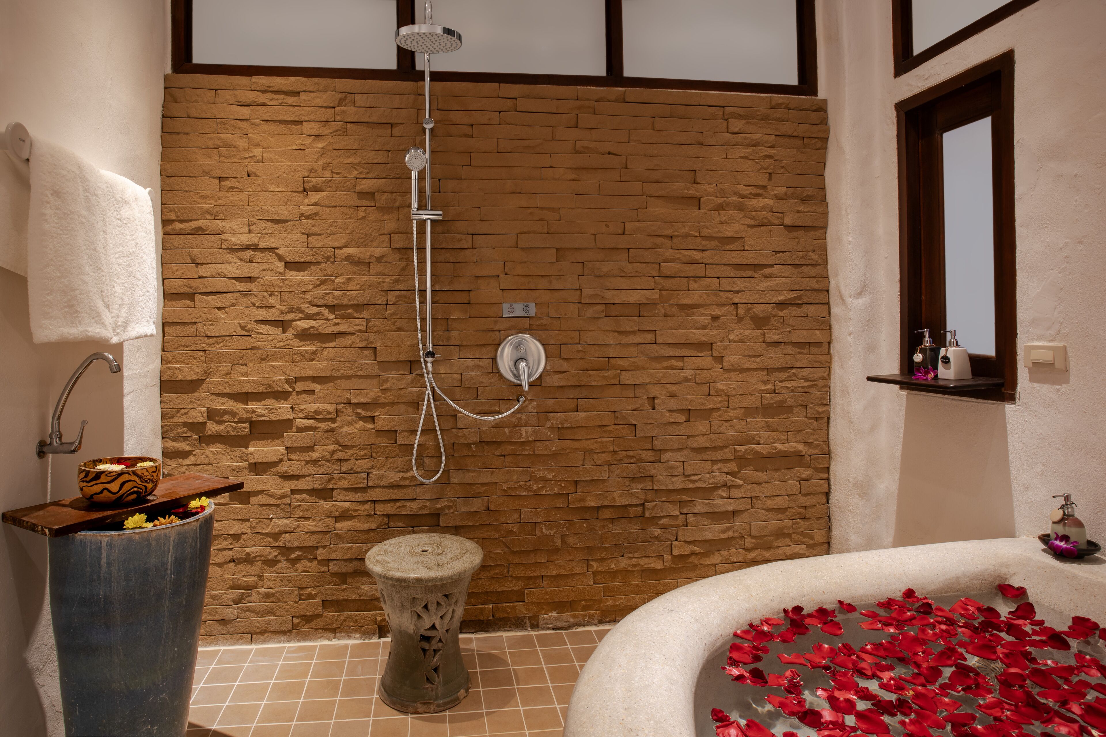 A bathroom with a stone wall, a shower, a bathtub with rose petals, a towel rack, and a small wooden stool.