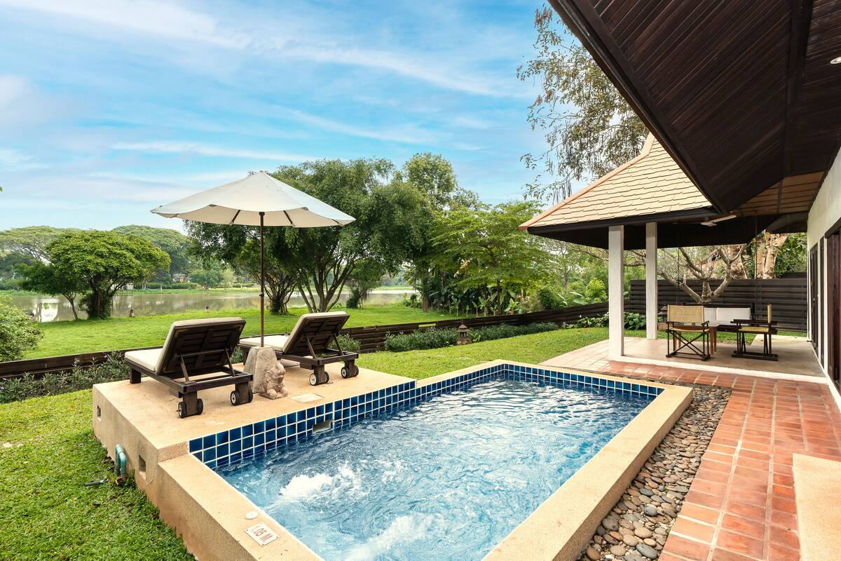 A rectangular swimming pool with a tiled edge, surrounded by a stone border. There are lounge chairs and a table with an umbrella in the background.