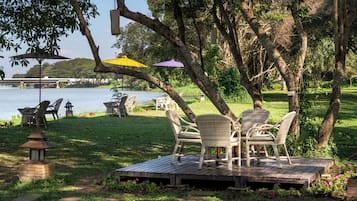 A deck with wicker chairs and a table, overlooking a lake with chairs and umbrellas in the background.