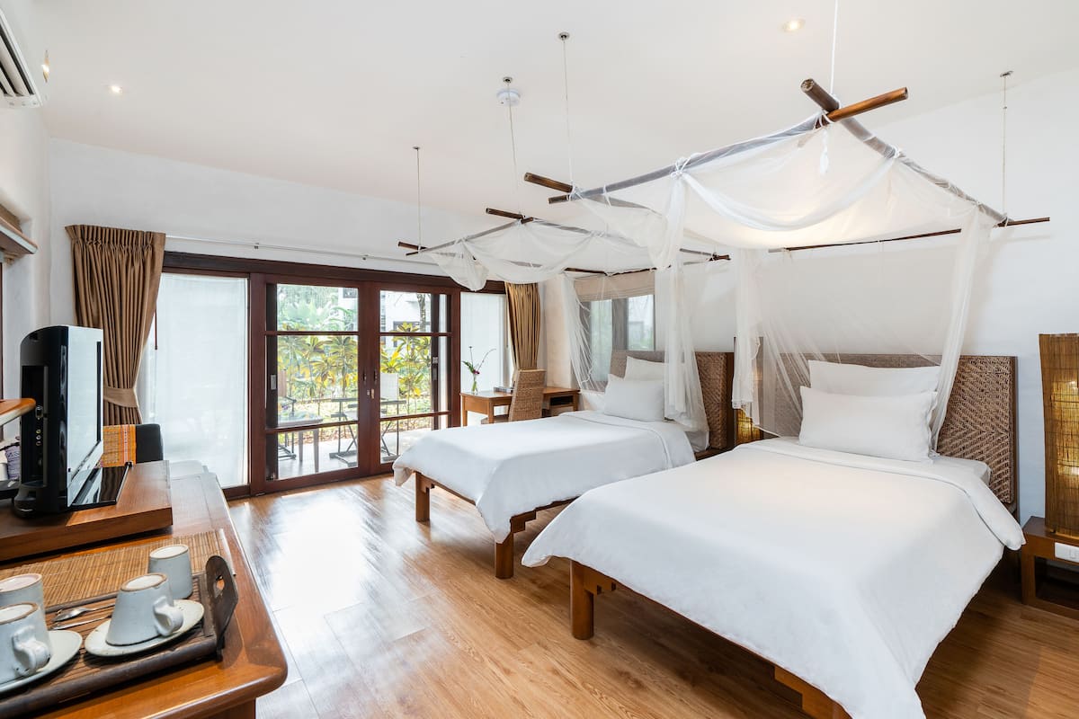 A hotel room with two beds, a wooden desk with a coffee maker, and a large window with curtains.