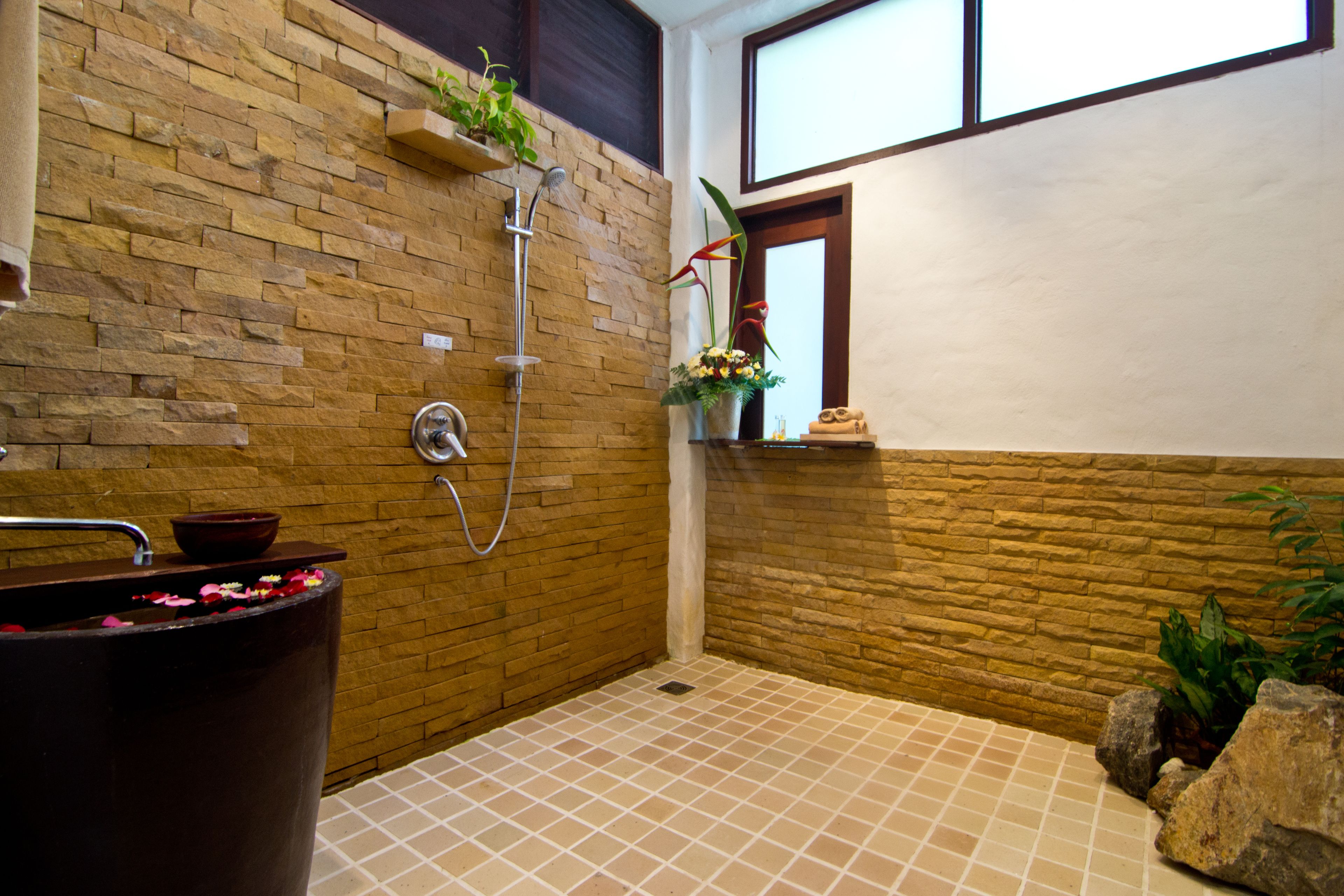 Ein Badezimmer mit einer Steinwand, einer Dusche, einer Badewanne mit Rosenblättern und einem Fenster mit Blick.