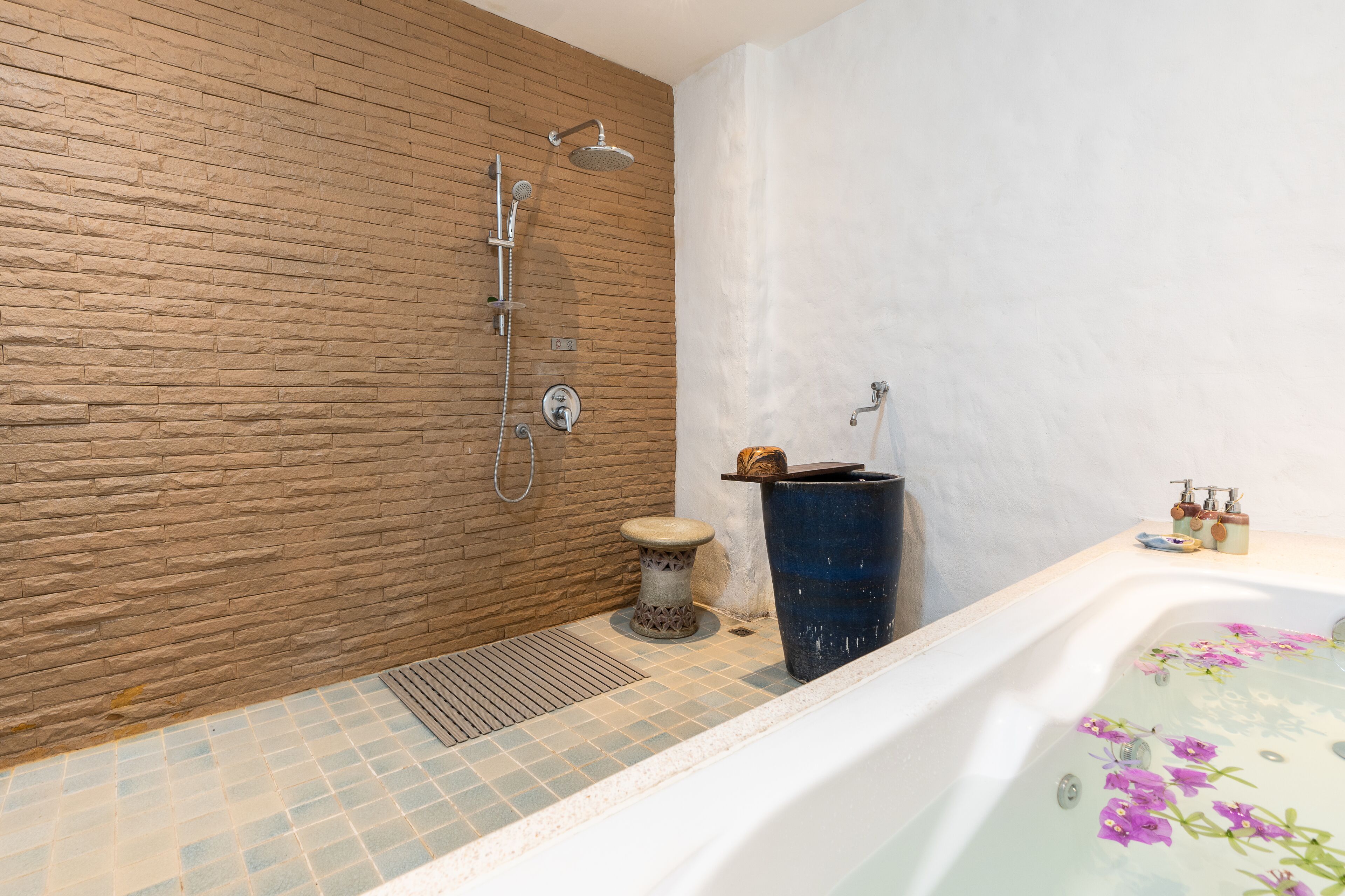 A bathroom with a stone wall, a shower, a bathtub with floral design, and a small stool.