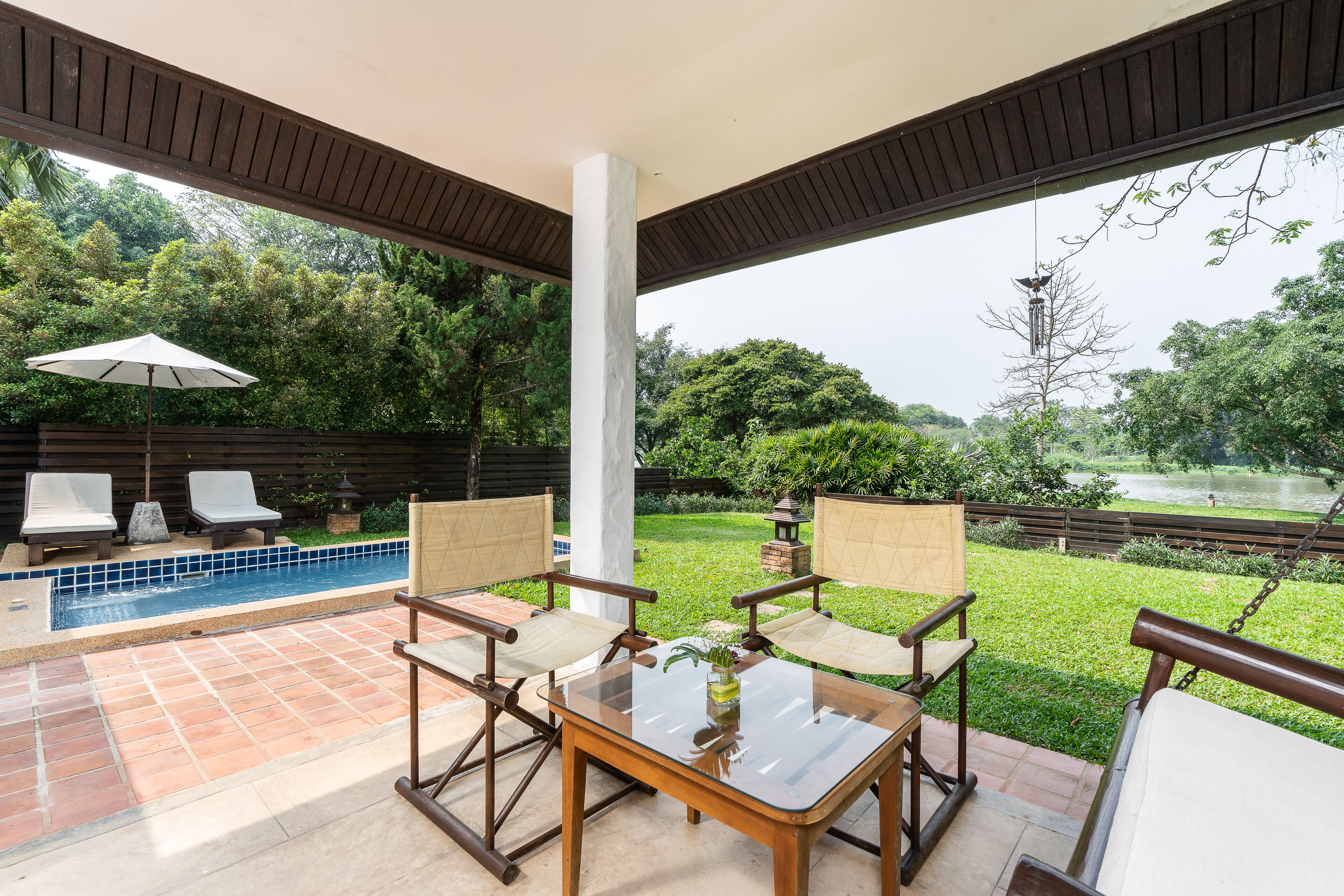 A covered patio area with a pool, two lounge chairs, a table with a vase, and a view of a pond and greenery.