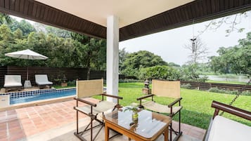 A covered patio area with a pool, two lounge chairs, a table with a vase, and a view of a pond and greenery.