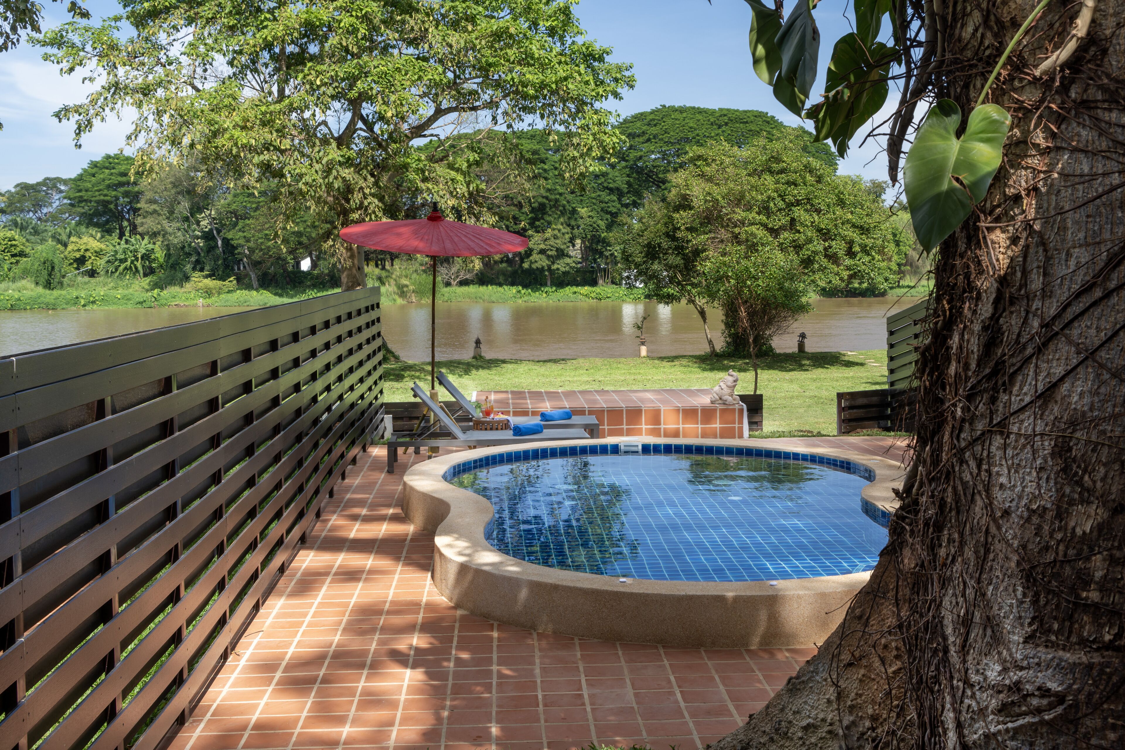 Une piscine dotée d’une terrasse en bois et d’un parasol rouge, entourée d’une végétation luxuriante et bordée par une rivière.