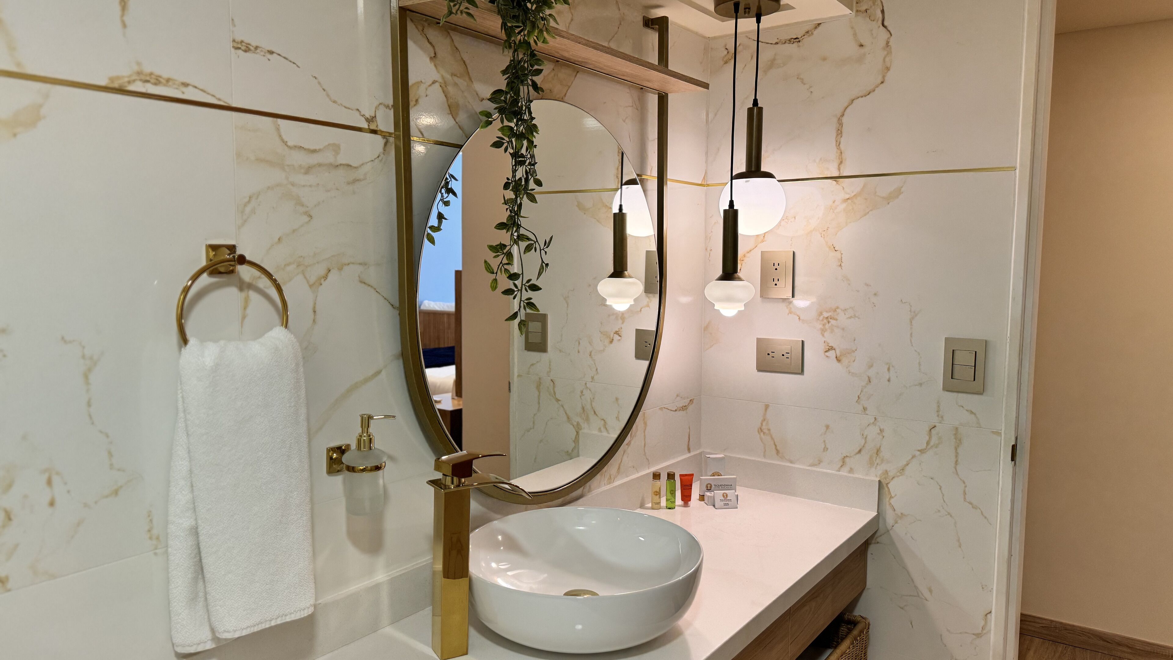 Ein Badezimmer mit Marmorwand, Waschbecken in Weiß, Wasserhahn in Gold, Handtuchhalter, Spiegel und einer hängenden Pflanze.