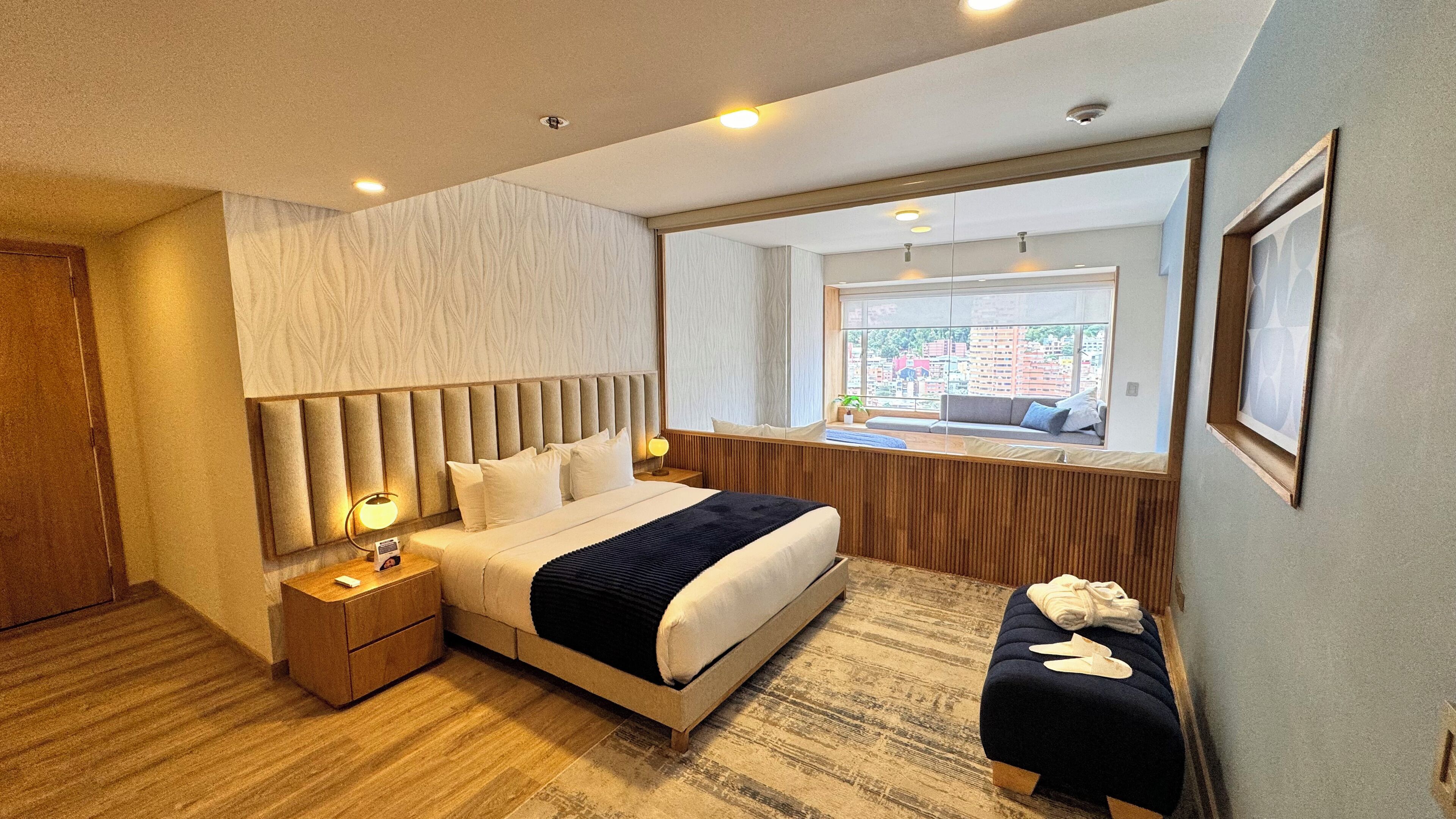 A modern hotel room with a large bed, a bedside table, a bench, and a window with a view of buildings.