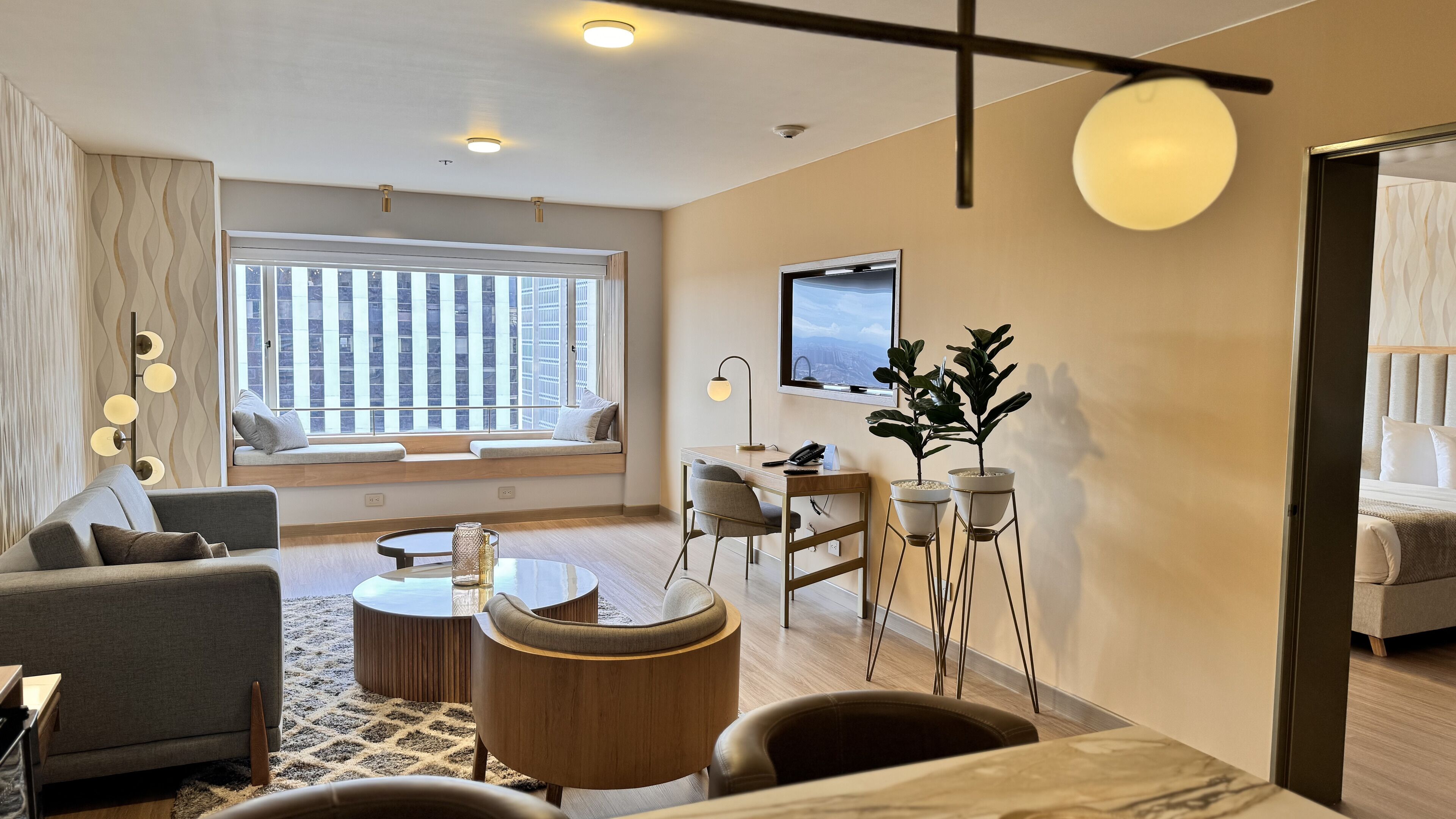 A modern hotel room with a sofa, a round coffee table, a desk with a chair, a TV mounted on the wall, and a window with a view of buildings.
