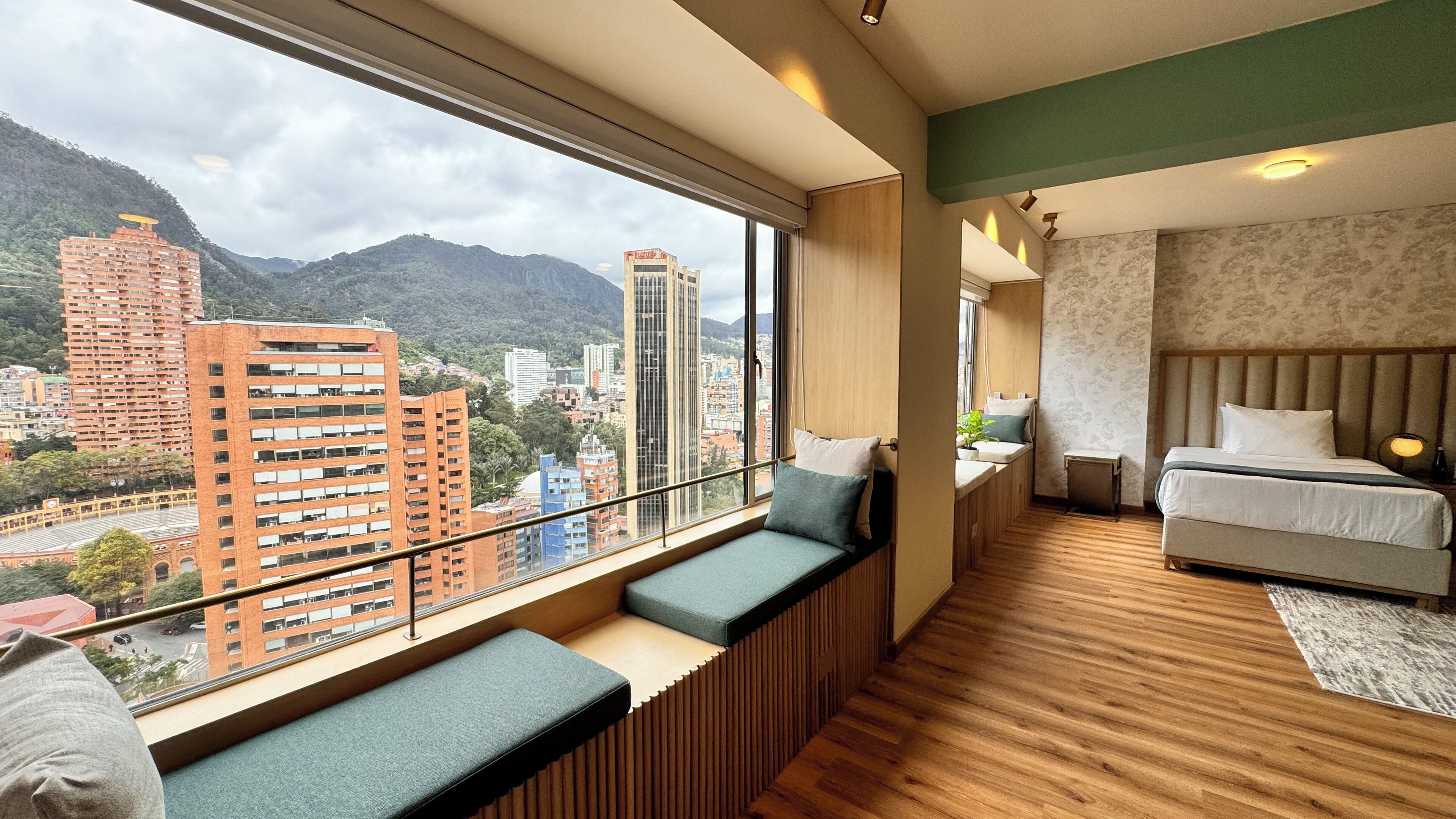 Una habitación de hotel moderna con un amplio ventanal que ofrece vistas a la ciudad, un rincón de descanso acogedor con cojines, una cama bien hecha y una mesita de noche con lámpara.