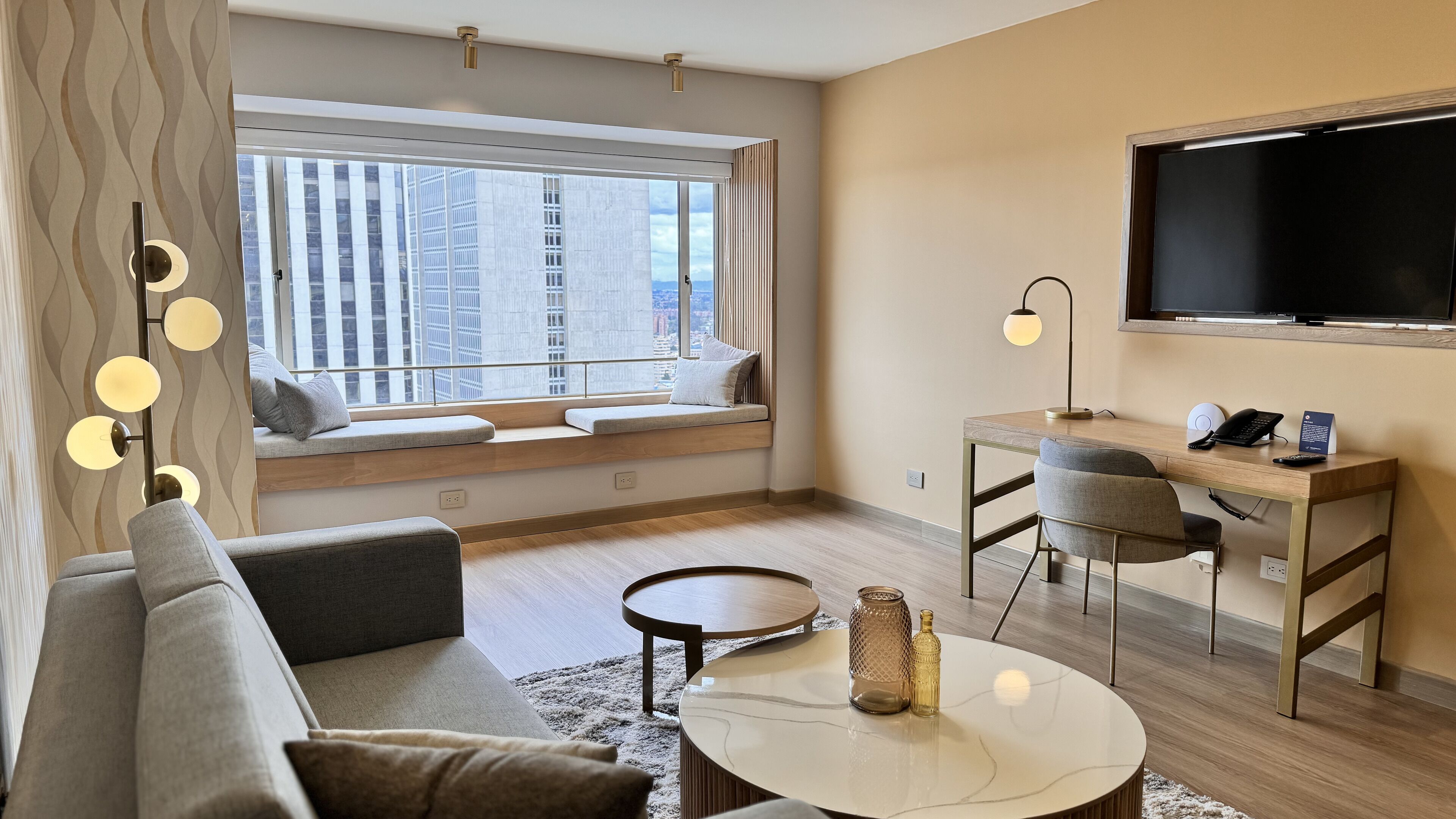 Un salon moderne comprenant un canapé, une table basse ronde, un bureau avec une chaise, un téléviseur fixé au mur et une grande fenêtre donnant sur la ville.