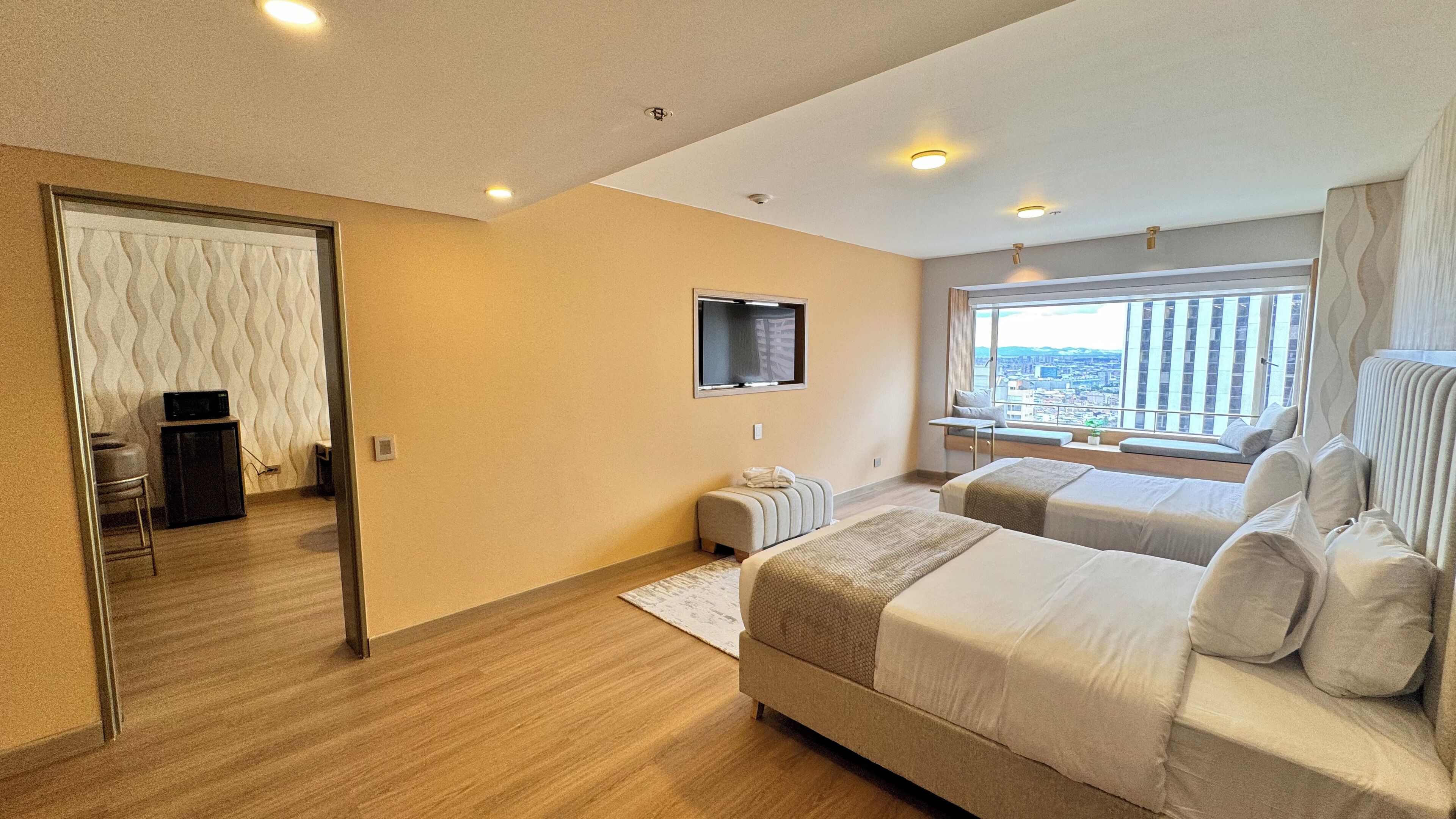 A modern hotel room with two beds, a TV, a window with a city view, and a small seating area by the window.
