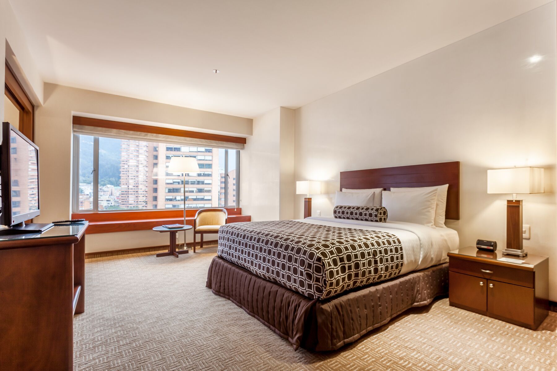 A hotel room with a large bed, a desk, a chair, and a window with a city view.