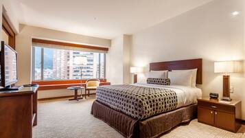 A hotel room with a large bed, a desk, a chair, and a window with a city view.