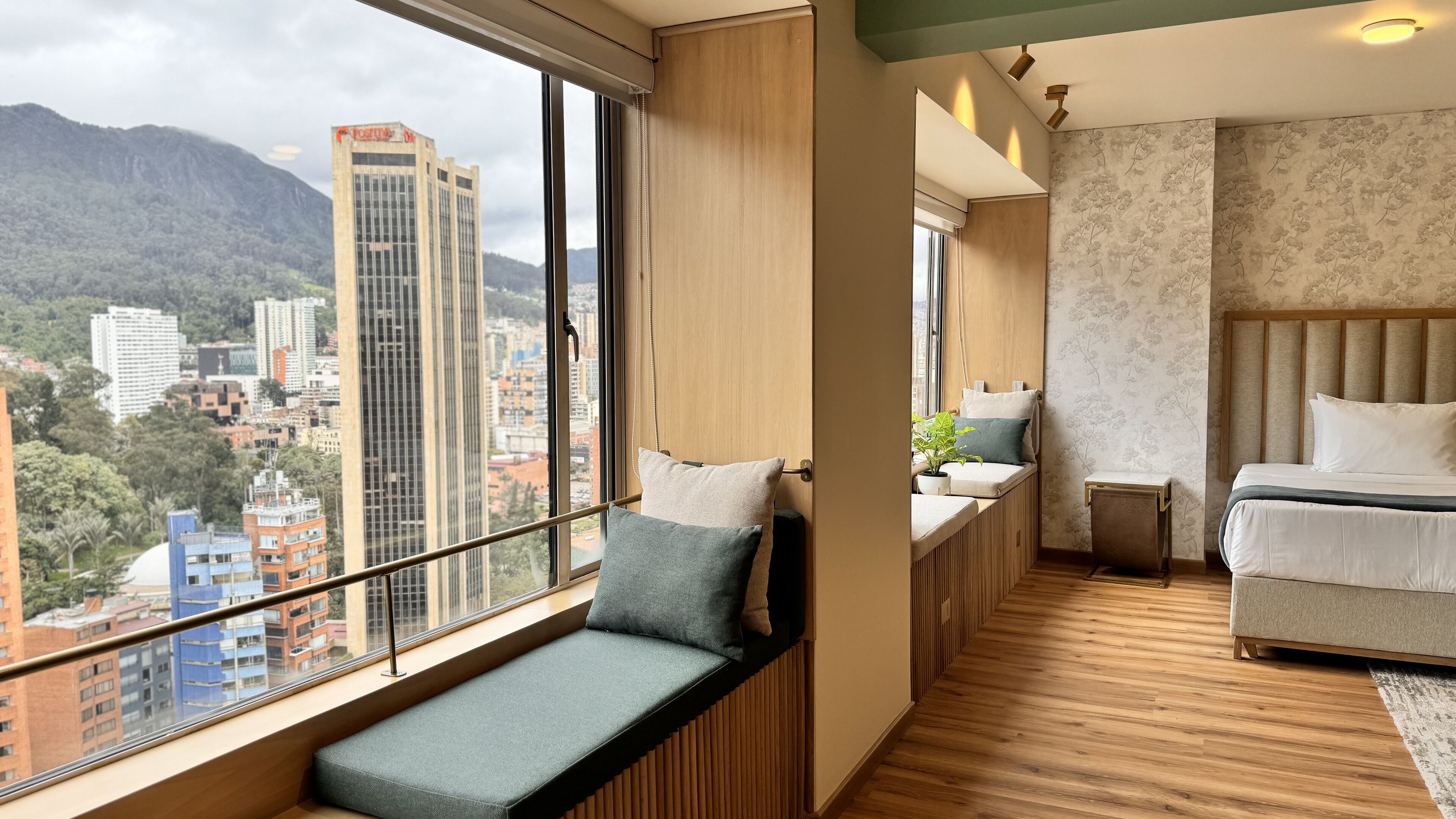 A modern hotel room with a large window offering a city view, a cozy seating area, a neatly made bed, and a small bedside table with a lamp.