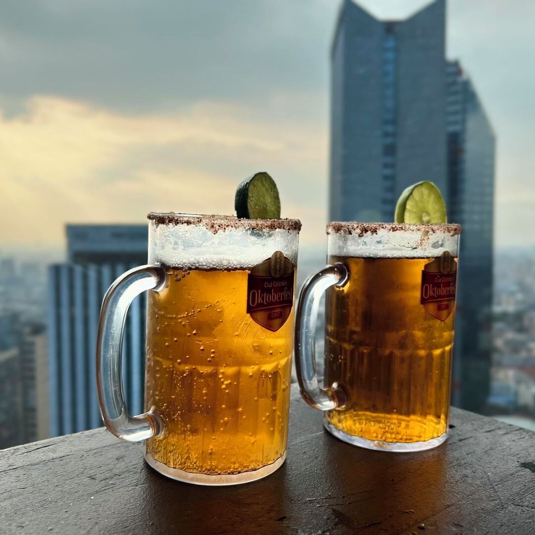 Two beer mugs with a frothy head and a lime slice, placed on a wooden surface with a cityscape and a tall building in the background.