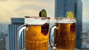 Two beer mugs with a frothy head and a lime slice, placed on a wooden surface with a cityscape and a tall building in the background.