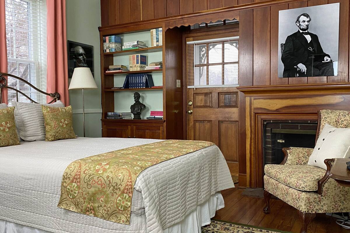 A bedroom with a large bed, a wooden bookshelf, a fireplace, and a portrait on the wall.