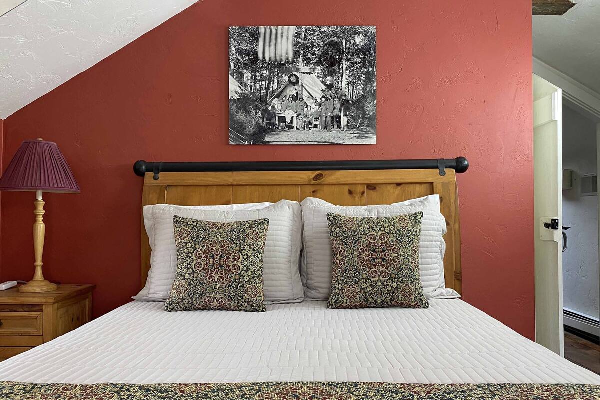 A bedroom with a wooden headboard, two patterned pillows, a white bedspread, a patterned rug, and a lamp on a wooden nightstand.