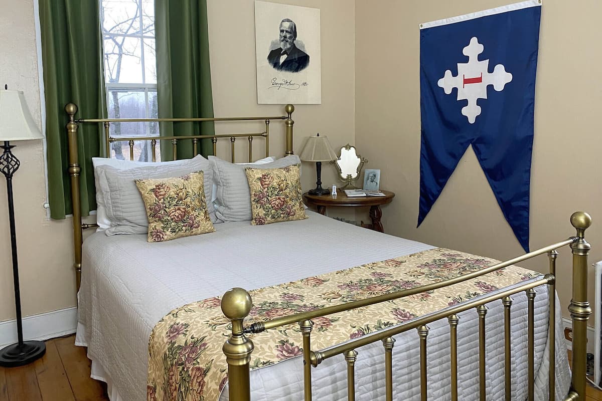 A neatly arranged bedroom with a bed, green curtains, a framed portrait, and a flag on the wall.