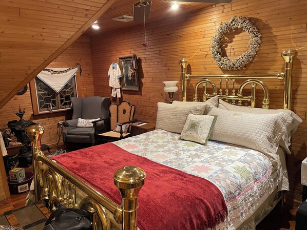 A cozy room with a wooden ceiling, a bed with a red blanket, a gold-framed headboard, a chair, a wall-mounted mirror, and a wreath.