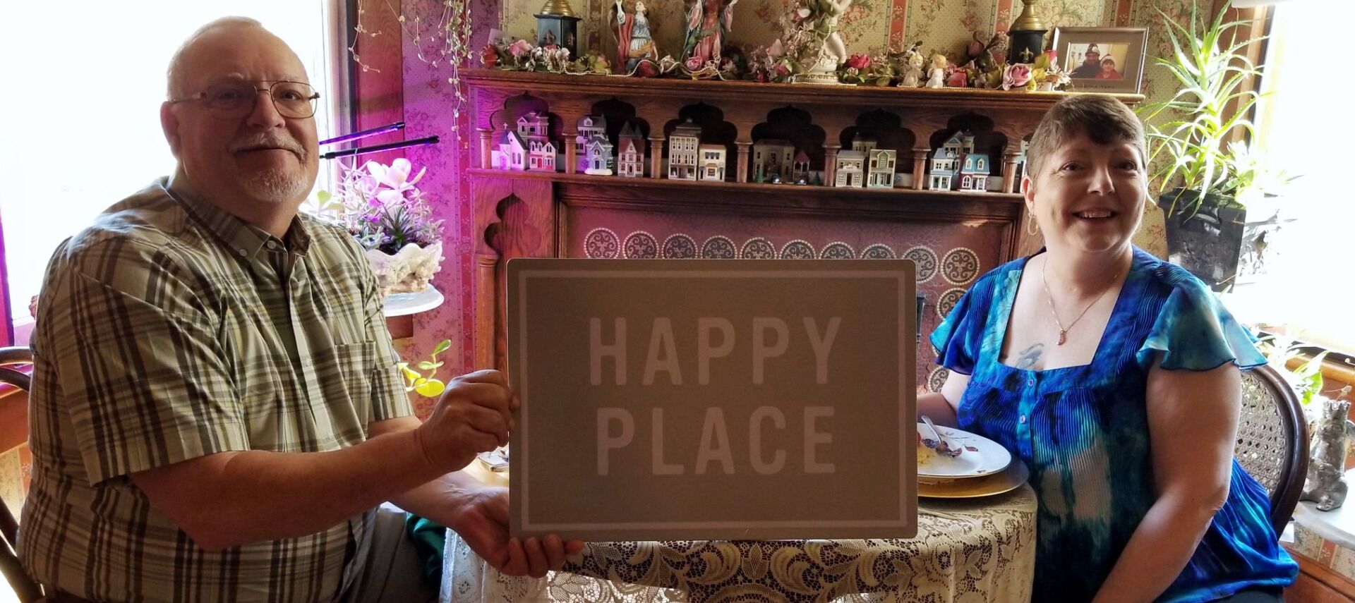 Dos personas sostienen un cartel que dice "HAPPY PLACE" en una habitación con un estante de madera que exhibe figuritas y una pequeña mesa con un plato y una taza.