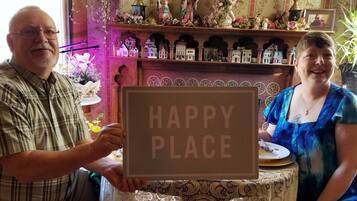 Dos personas sostienen un cartel que dice "HAPPY PLACE" en una habitación con un estante de madera que exhibe figuritas y una pequeña mesa con un plato y una taza.