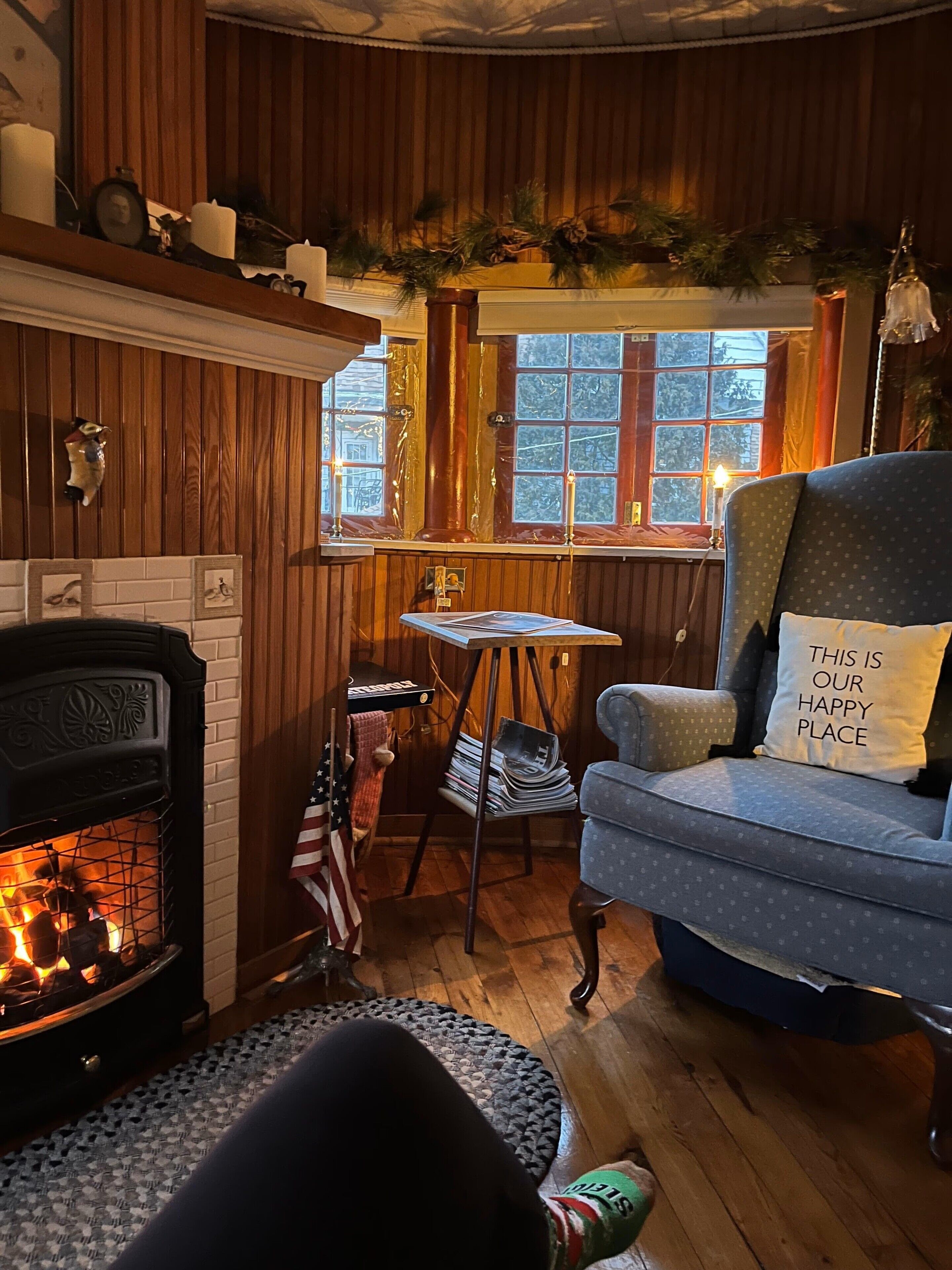A cozy room with a fireplace, a blue armchair with a pillow, a small table, and a window with a view.