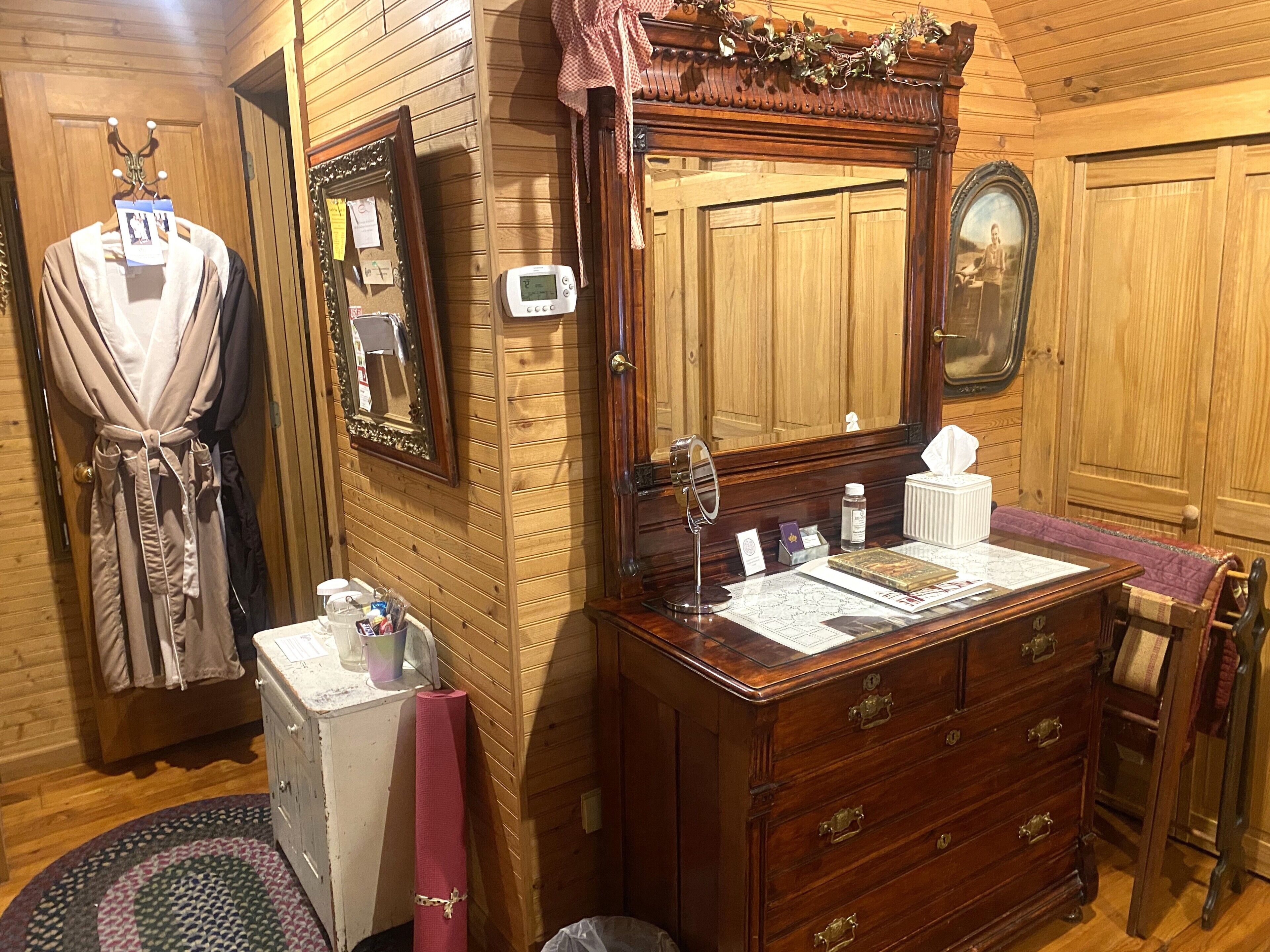 A wooden-paneled room with a vanity, a mirror, a robe, and a small table with toiletries.