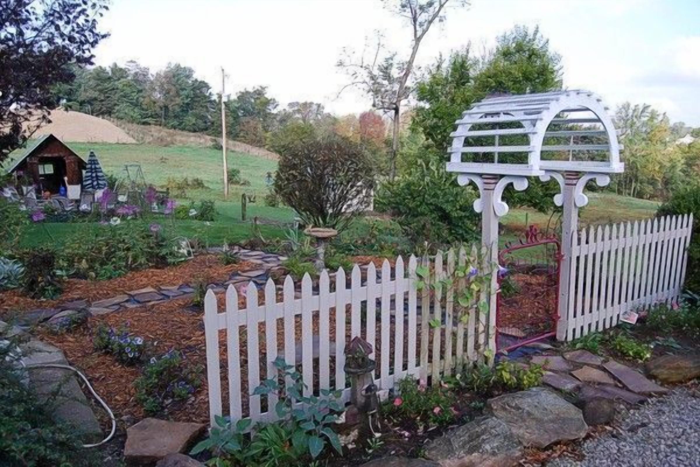 Un jardin avec une clôture blanche en bois, une petite construction en bois et une pergola blanche.