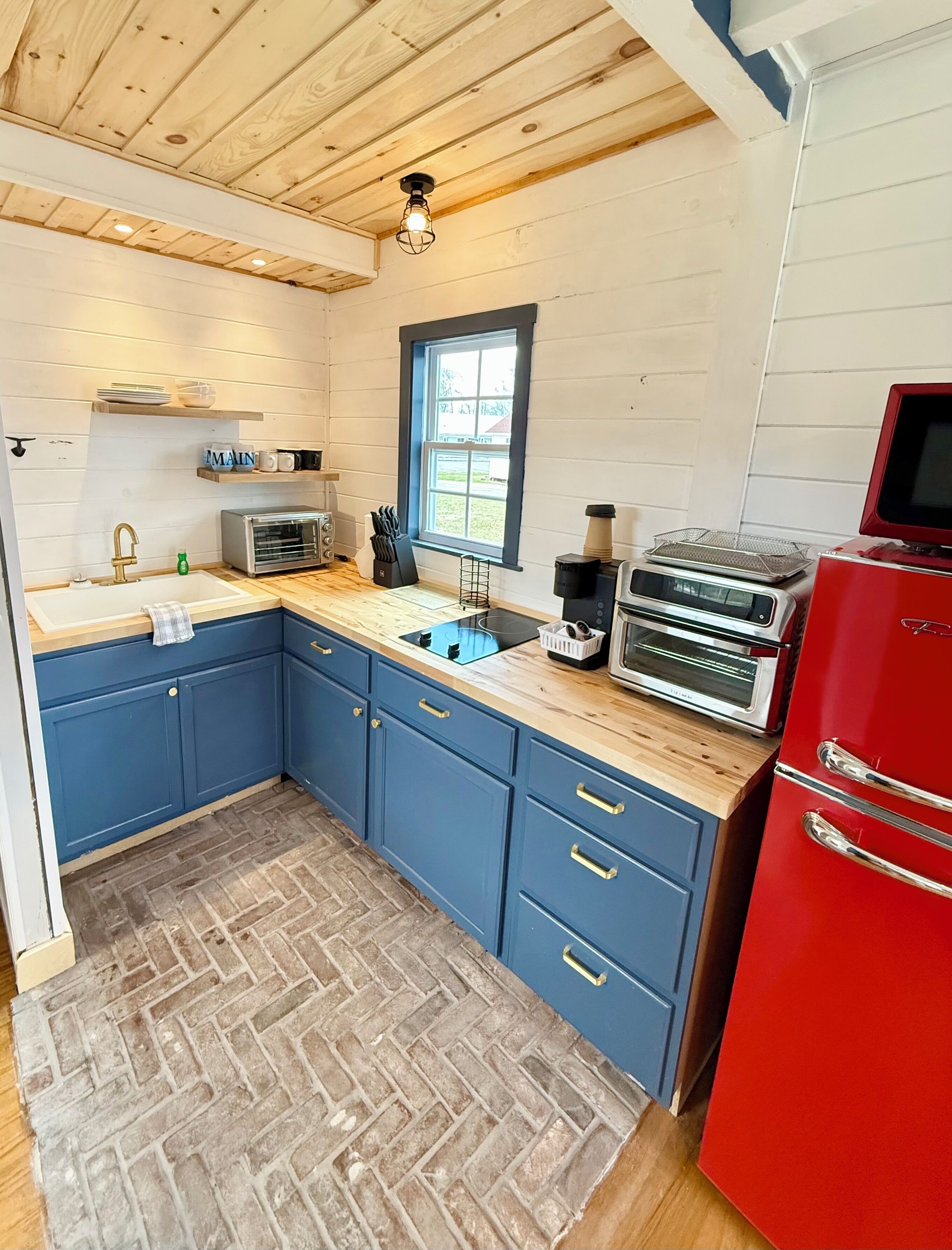 A kitchen with blue cabinets, a red refrigerator, a window, and a wooden ceiling.