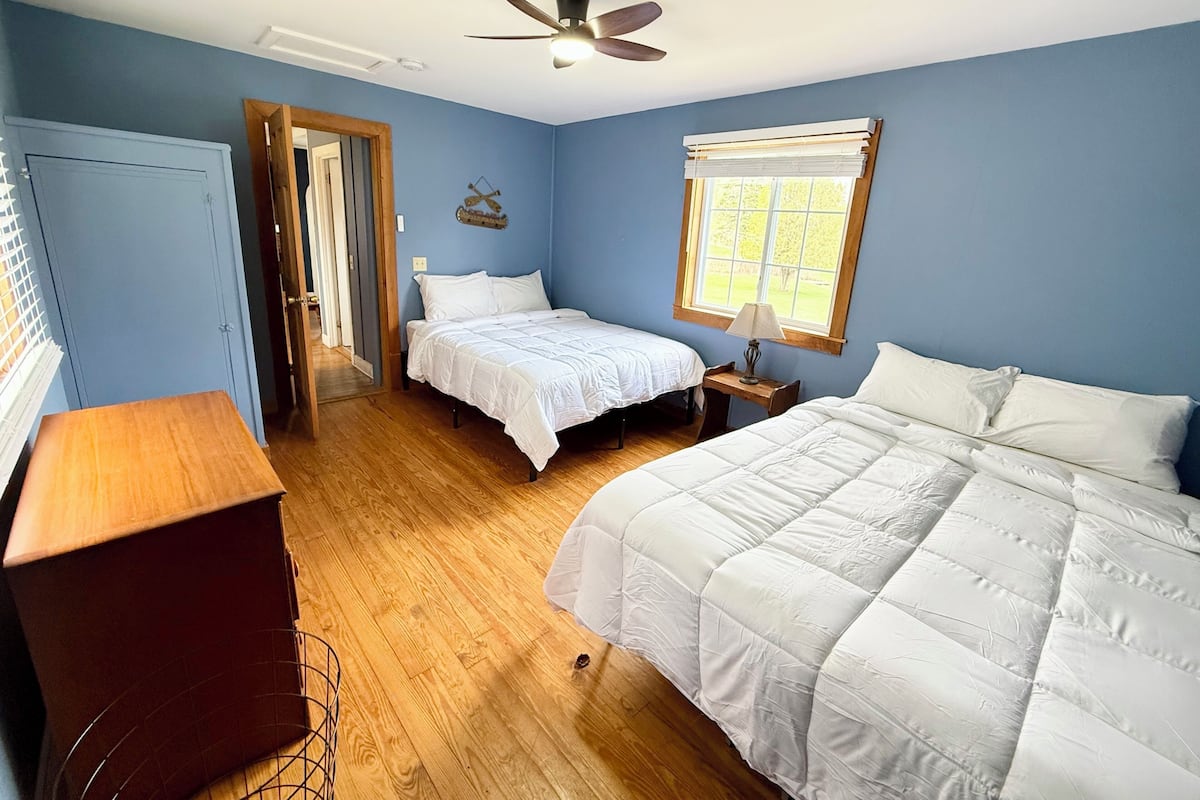 A bedroom with two beds, a ceiling fan, a window with blinds, and a wooden floor.