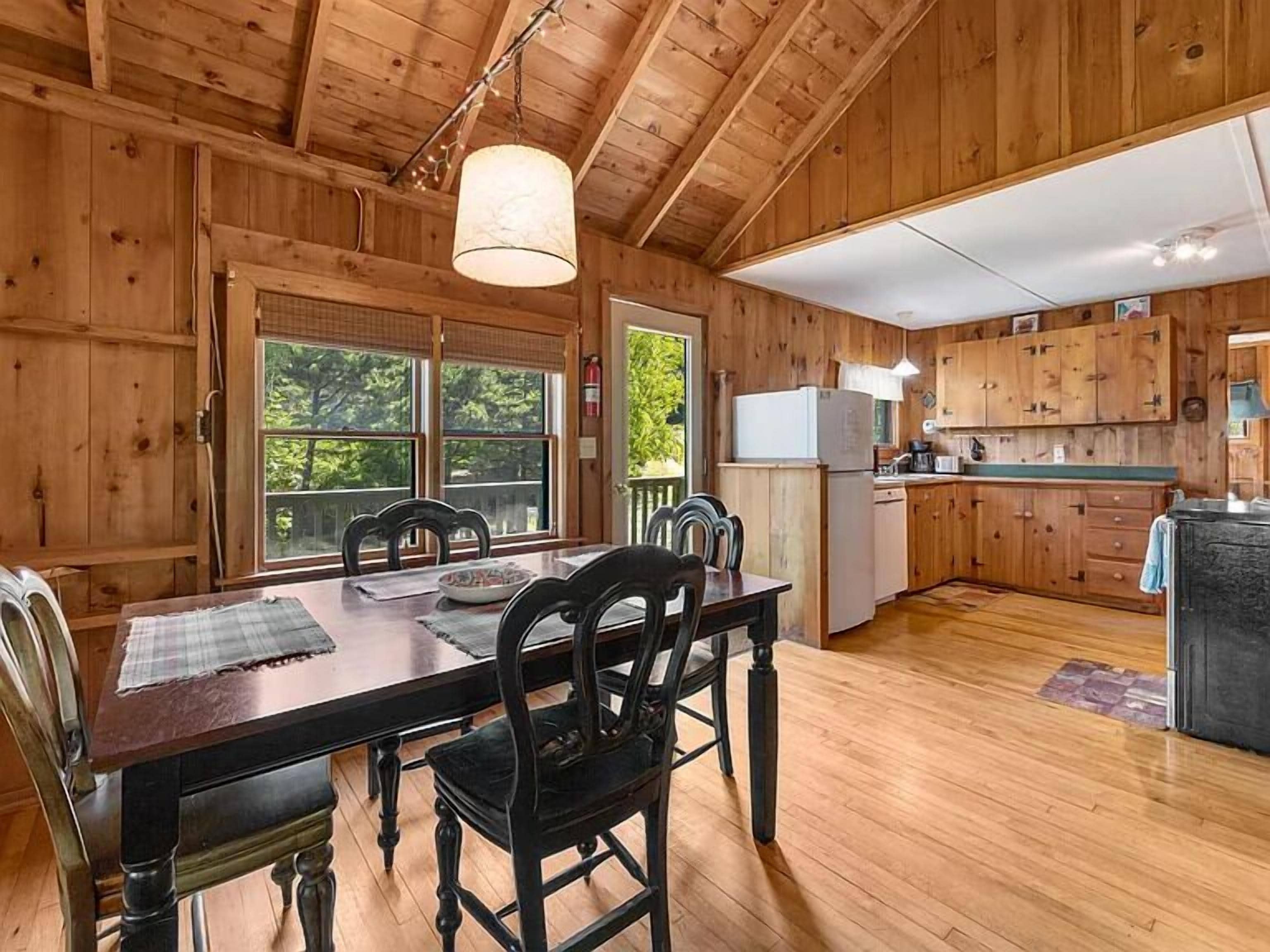 A wooden cabin interior with a dining table, chairs, a kitchen area, and large windows.