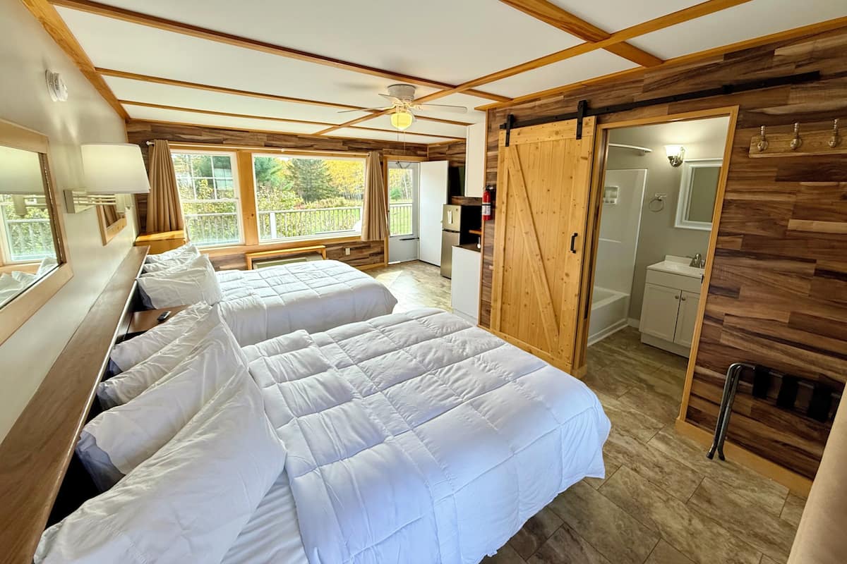 A room with two beds, a ceiling fan, a sliding door to a bathroom, and a large window with a view of trees.
