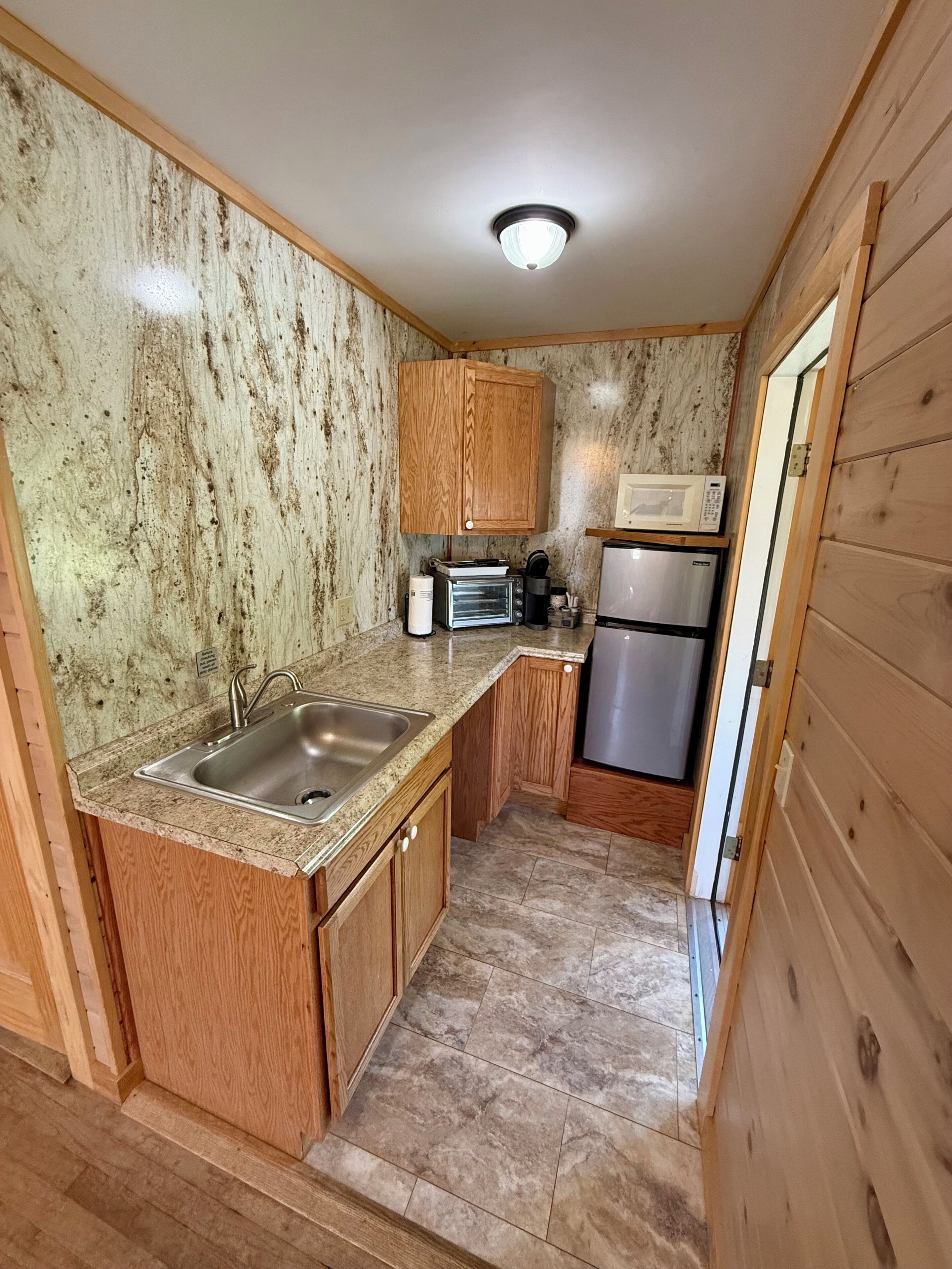 A compact kitchen with wooden cabinets, a stainless steel sink, a microwave, a toaster, and a refrigerator.