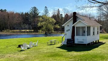 A small white cabin with a chimney, situated on stilts by a lake. There are picnic tables and chairs on the grass in front.