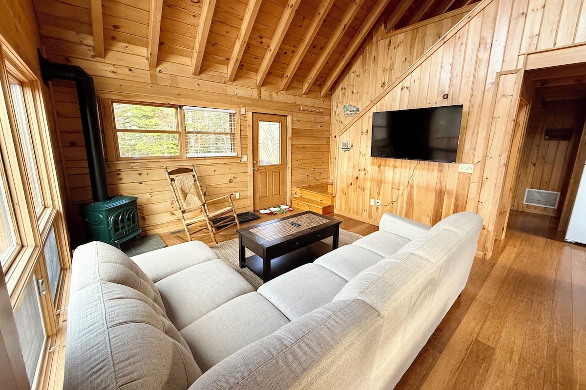 A cozy cabin interior with a sofa, a wooden ceiling, a television, and a fireplace.