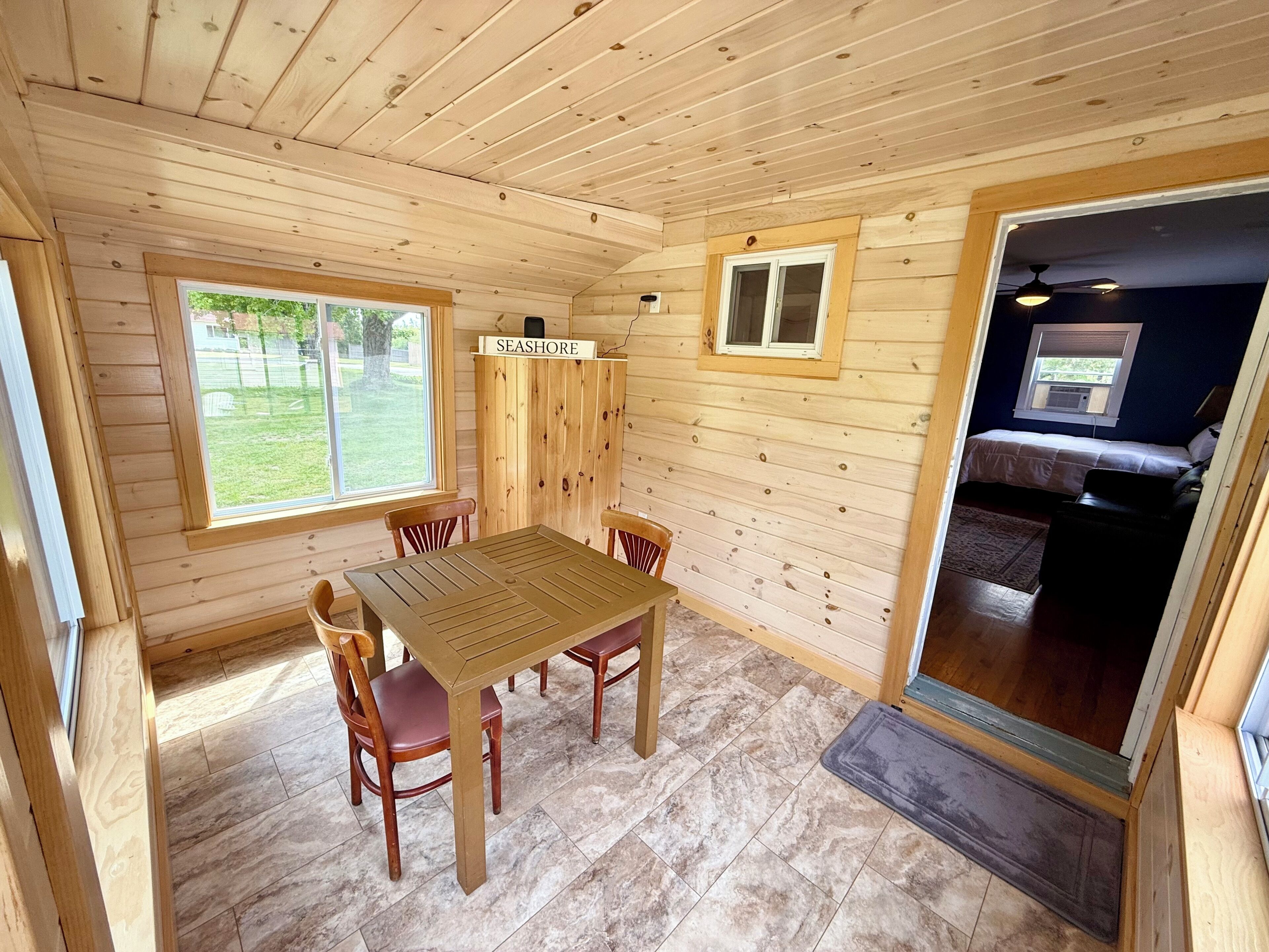 A wooden interior with a dining table and chairs, a window with a view of a grassy area, and a sign labeled 'SEASHORE'.