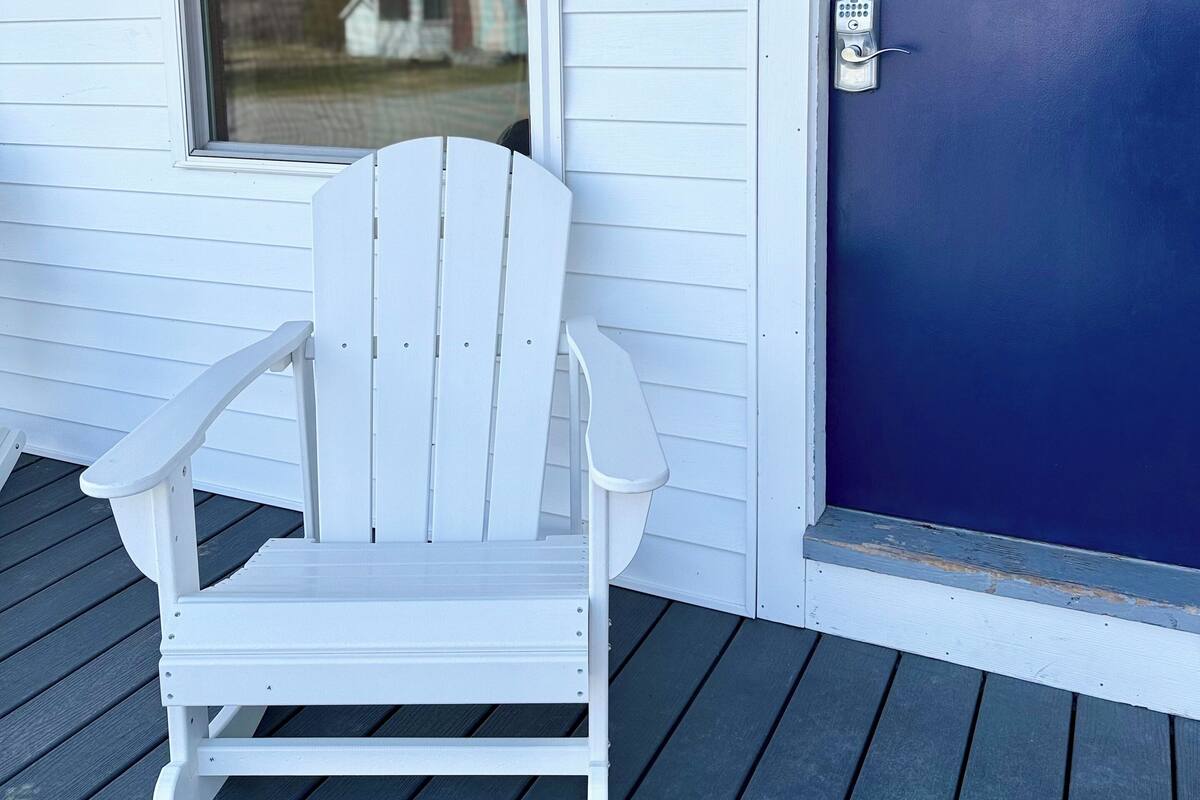 A white Adirondack chair on a wooden deck, a blue door, and a window with a view of houses.