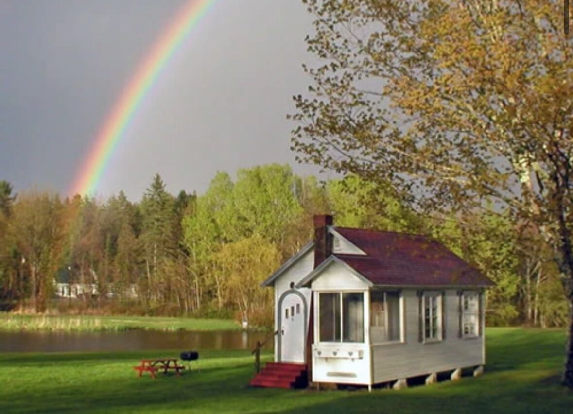 A small white cabin with a porch, situated by a lake with a rainbow in the background.