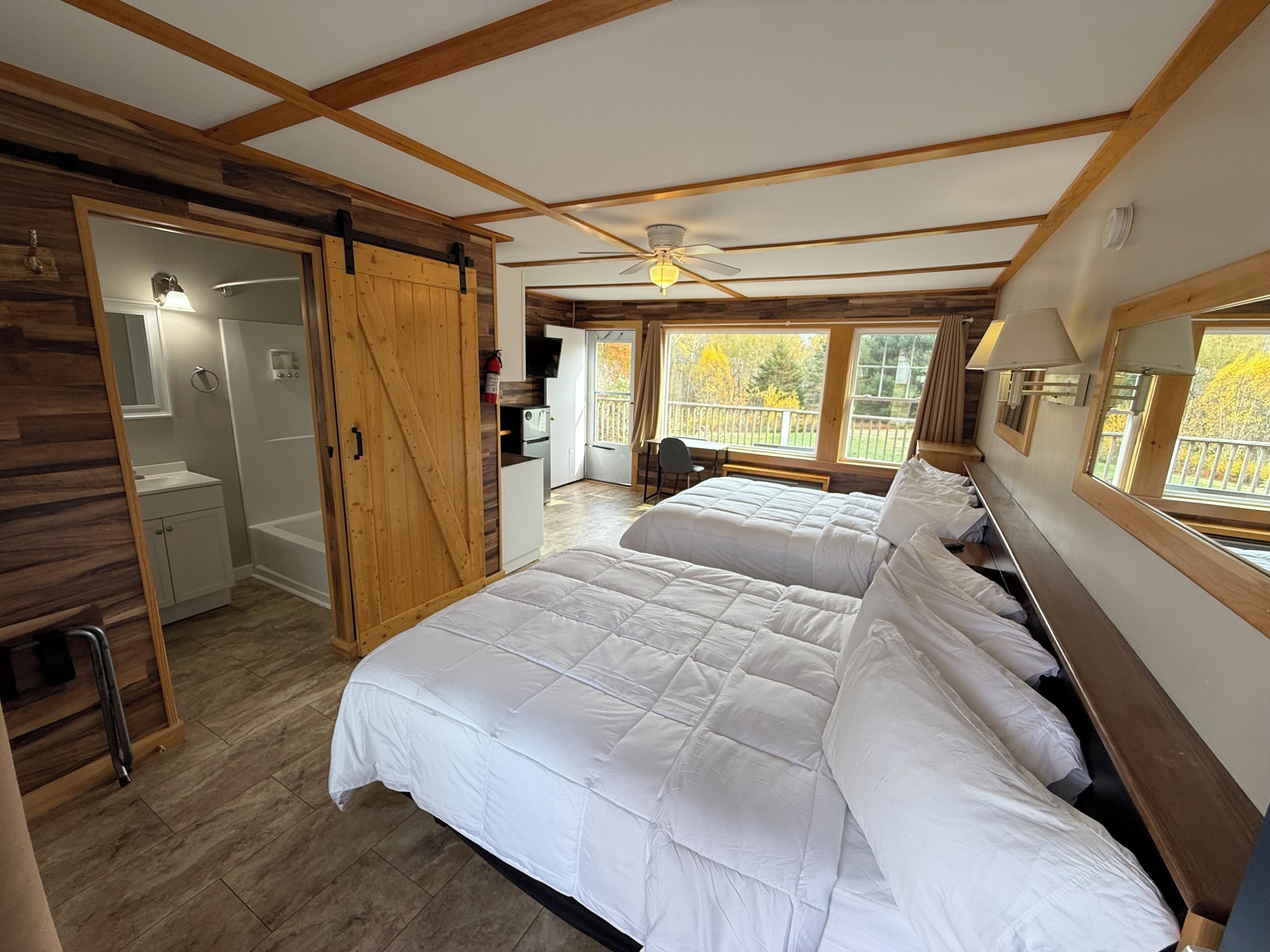 A bedroom with two beds, a wooden wardrobe, a bathroom, and a balcony with a view of trees.