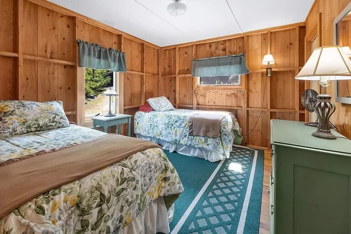 A room with two beds, a window with curtains, a lamp, and a fan.