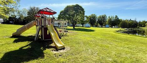 A playground with a slide and swing set, surrounded by a grassy field and trees.