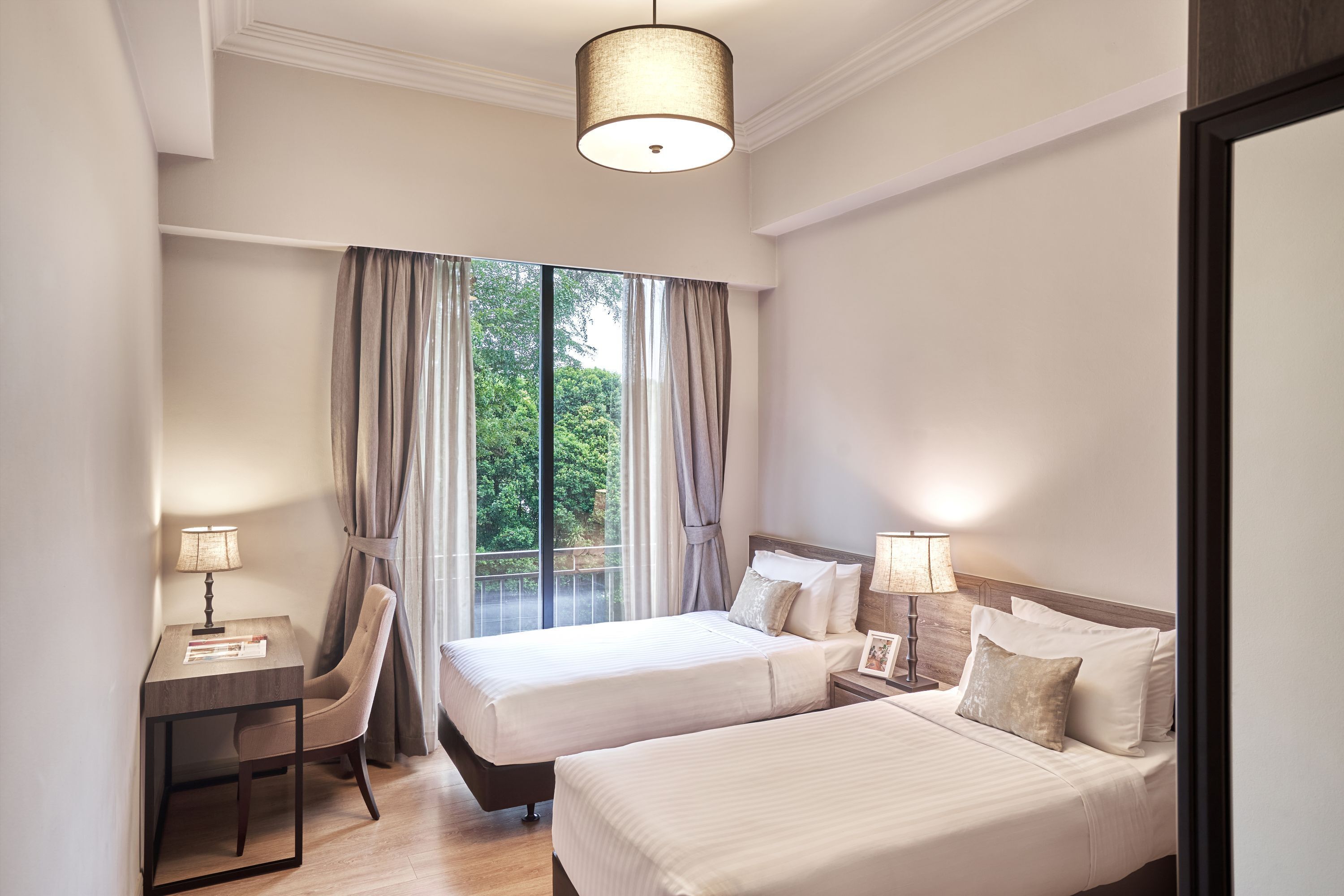 A hotel room with two beds, a desk, a chair, and a view of greenery through the window.