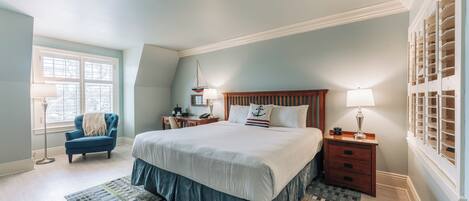 A bedroom with a large bed, a chair, a nightstand, a lamp, and a window with blinds.