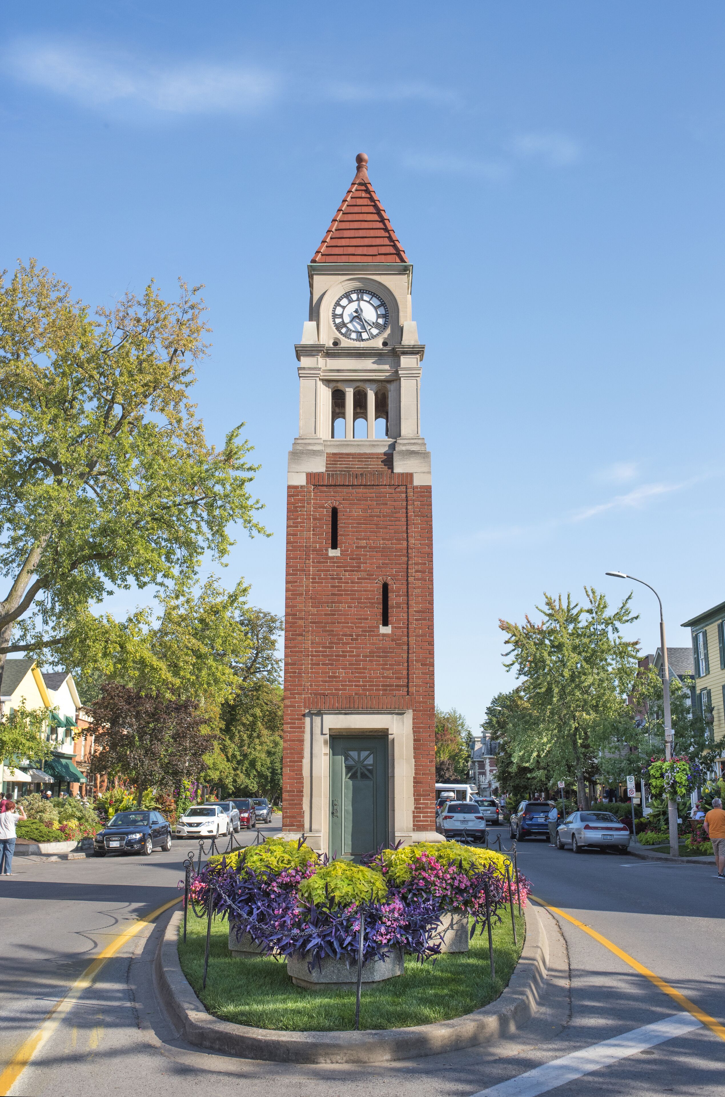 Una torre del reloj con una puerta verde, rodeada por un parterre de flores moradas.
