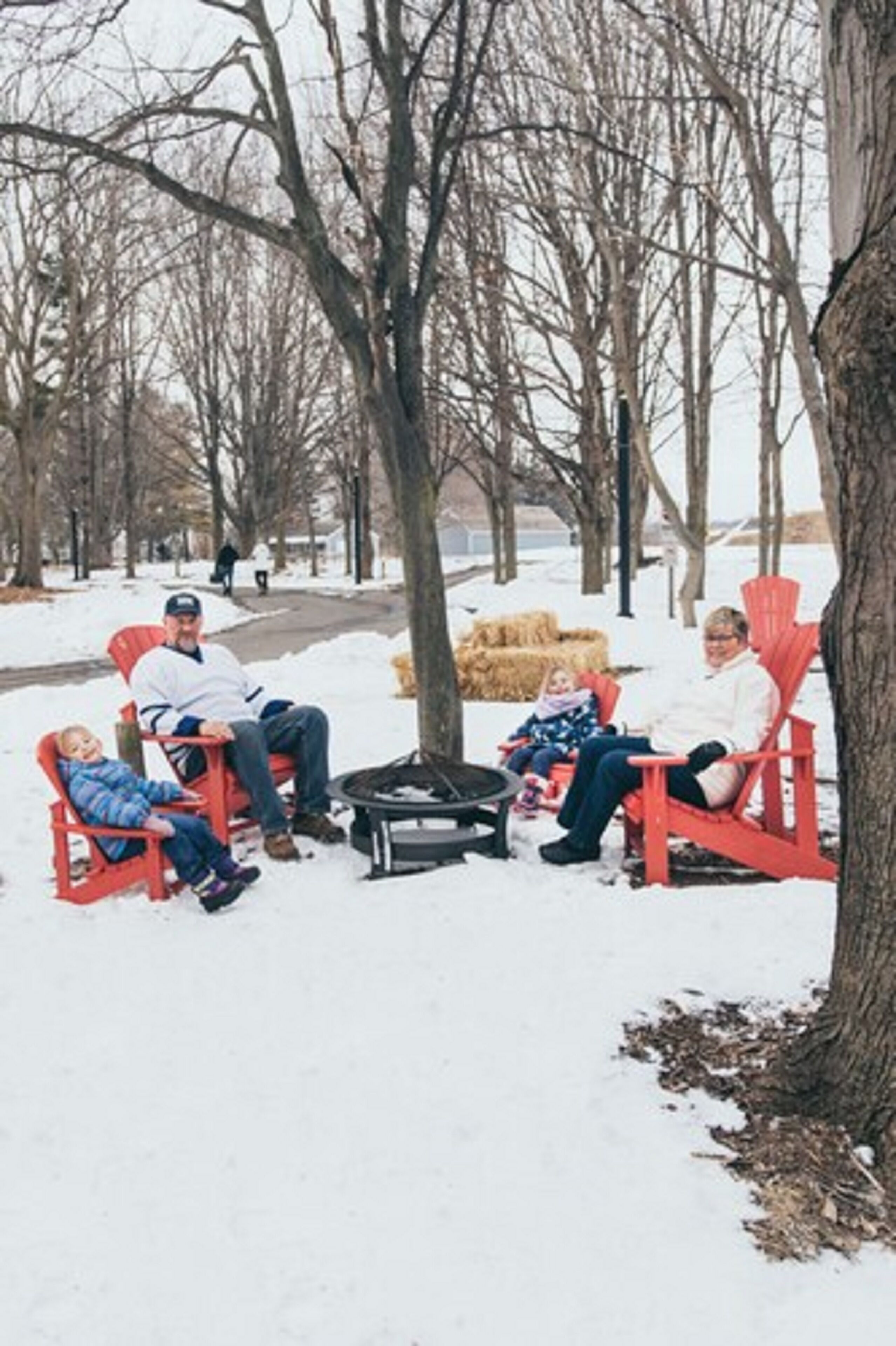 Un grupo de personas sentadas en sillones Adirondack rojos en un parque nevado, con árboles desprovistos de hojas y fardos de heno al fondo.